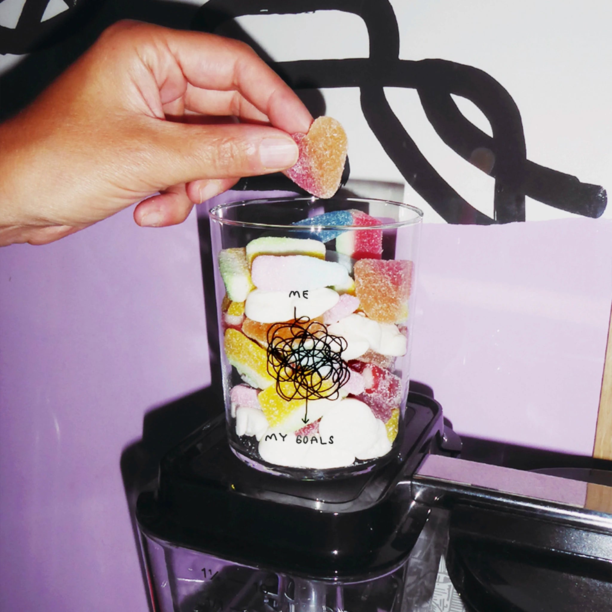 Transparentes großes Designer Glas mit schwarzem handschriftlichen Druck und Illustration "ME AND MY GOALS". Zu sehen ist die Verbindung von ME und MY GOALS wie diese wirr in hektischen Linien mit vielen Kurven und Kreisen verbunden werden. 
Das Glas hat sehr dünne, filigrane Wände und wird von zwei Händen gehalten. Das Glas ist mit verschiedenen Süßigkeiten befüllt.