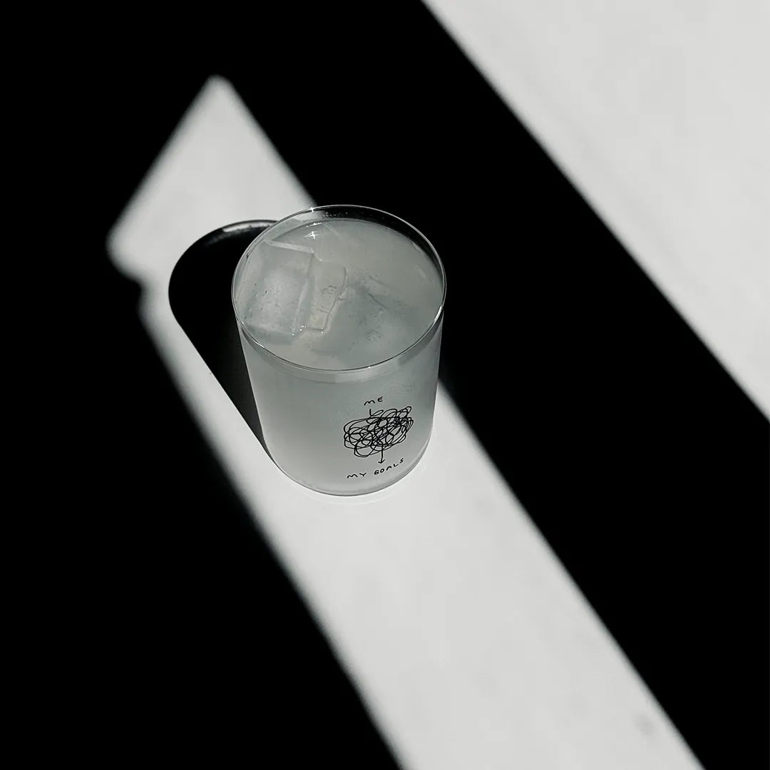 Transparentes Medium Designer Glas mit schwarzem Schriftzug "me" und "my goals". Dazwischen ist eine sehr hektische Linie mit vielen Kreisen, die beschreibt, dass der Weg zwischen "me" and "my goals" sehr komplex und hektisch ist. Das Glas hat dünne Wände und Boden. Das Glas steht im Licht auf einem weißen Tisch. Der Rest des Bildes ist dunkel in Schatten gefallen.

Das Glas ist befüllt mit einer klaren Flüssigkeit und einem Eiswürfel.
