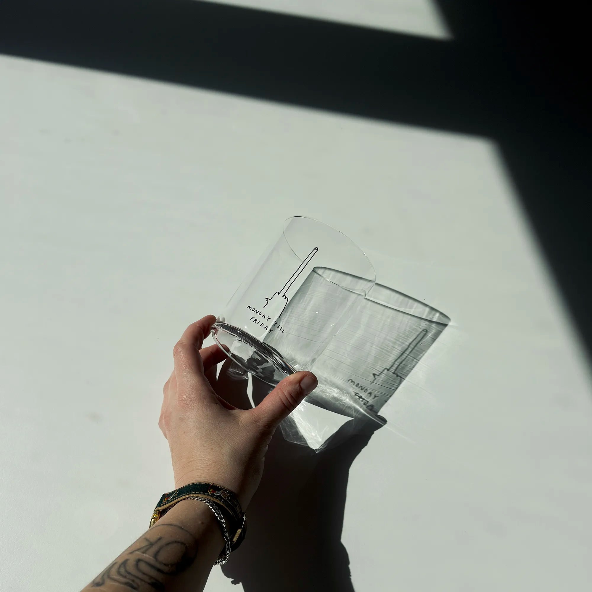 Großes Designer-Trinkglas mit sehr dünnen Glaswänden und einer schwarzen, einfarbigen Illustration auf der Vorderseite mit dem Motiv „Monday till Friday“. Illustration von Künstlerin, Illustratorin und Co-Gründerin Johanna Schwarzer.

Das Glas steht auf einem weißen Tisch, ist ungefüllt und liegt. Dabei wird es in der Hand gehalten, man sieht ein Tattoo und eine Kette. 