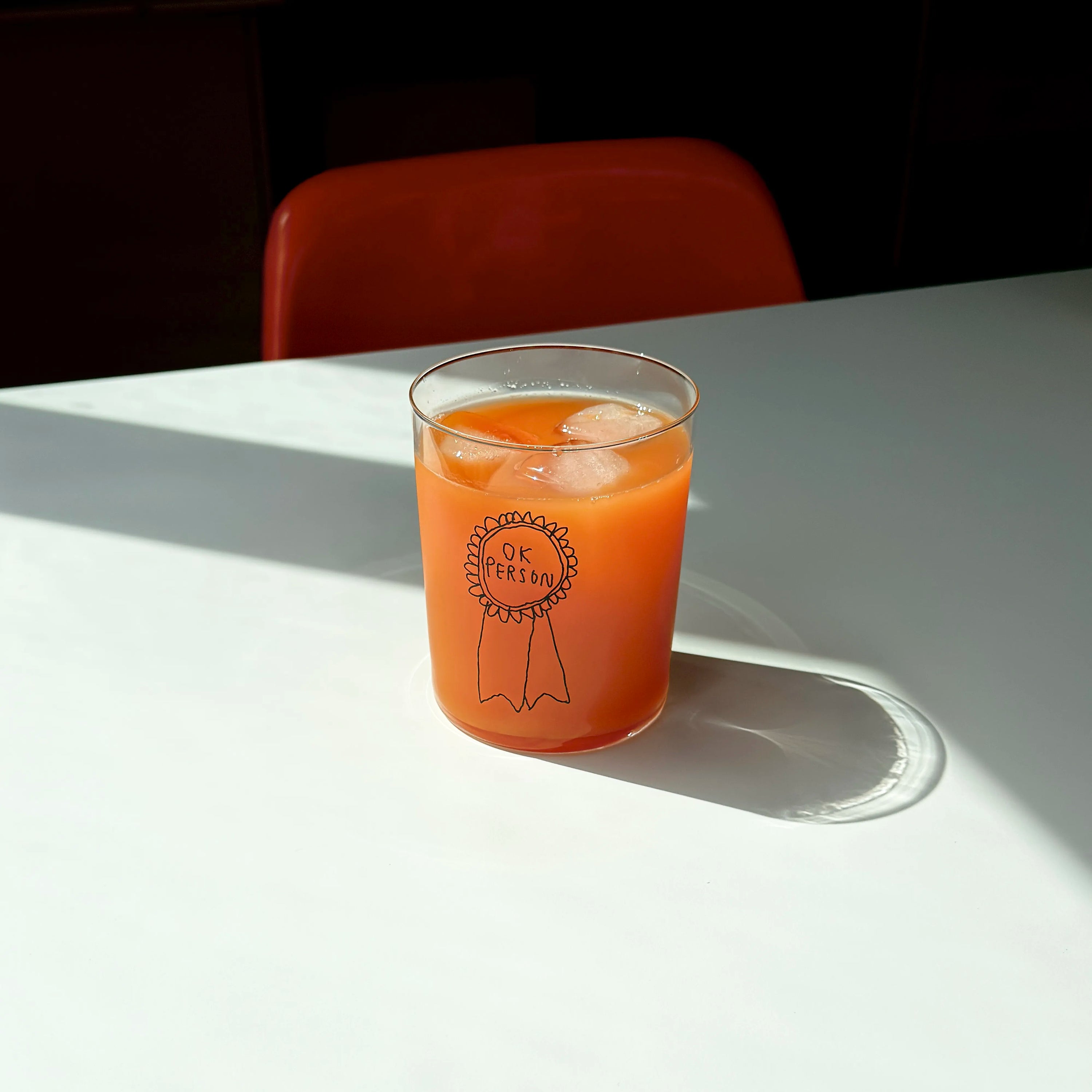 Klares, großes Designer-Glas mit sehr dünnen Wänden und Boden. Auf der Vorderseite ist eine schwarze, einfarbige Illustration einer Brosche abgebildet, auf der „ok person“ steht. Die Illustration wurde von der Künstlerin und Illustratorin Johanna Schwarzer gestaltet.
Das Glas steht auf einem weißen Tisch und ist mit einer orangenen Flüssigkeit befüllt. 