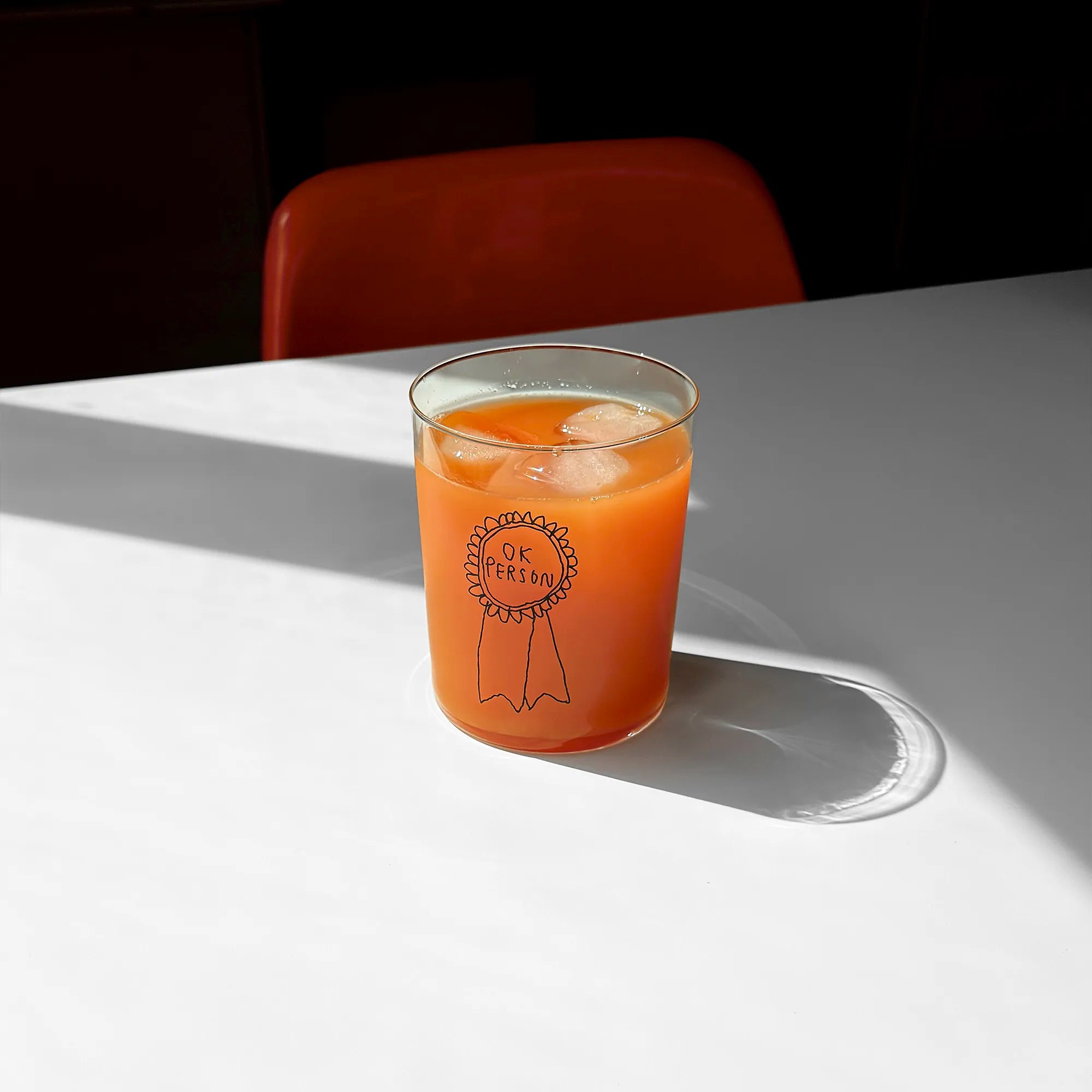 Klares, großes Designer-Glas mit sehr dünnen Wänden und Boden. Auf der Vorderseite ist eine schwarze, einfarbige Illustration einer Brosche abgebildet, auf der „ok person“ steht. Die Illustration wurde von der Künstlerin und Illustratorin Johanna Schwarzer gestaltet.
Das Glas steht auf einem weißen Tisch und ist mit einer orangenen Flüssigkeit befüllt. 