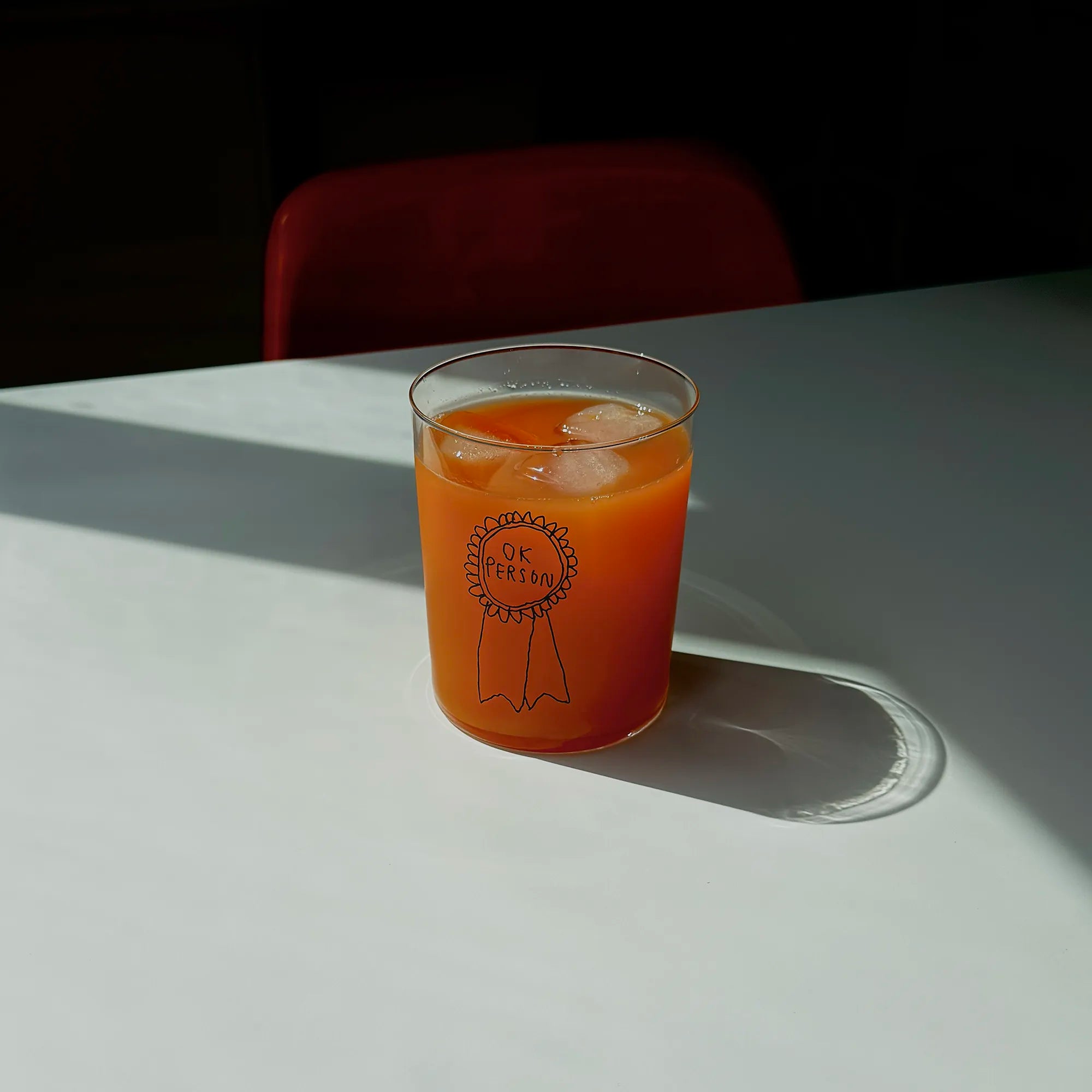 Klares, großes Designer-Glas mit sehr dünnen Wänden und Boden. Auf der Vorderseite ist eine schwarze, einfarbige Illustration einer Brosche abgebildet, auf der „ok person“ steht. Die Illustration wurde von der Künstlerin und Illustratorin Johanna Schwarzer gestaltet.
Das Glas steht auf einem weißen Tisch und ist mit einer orangenen Flüssigkeit befüllt. 