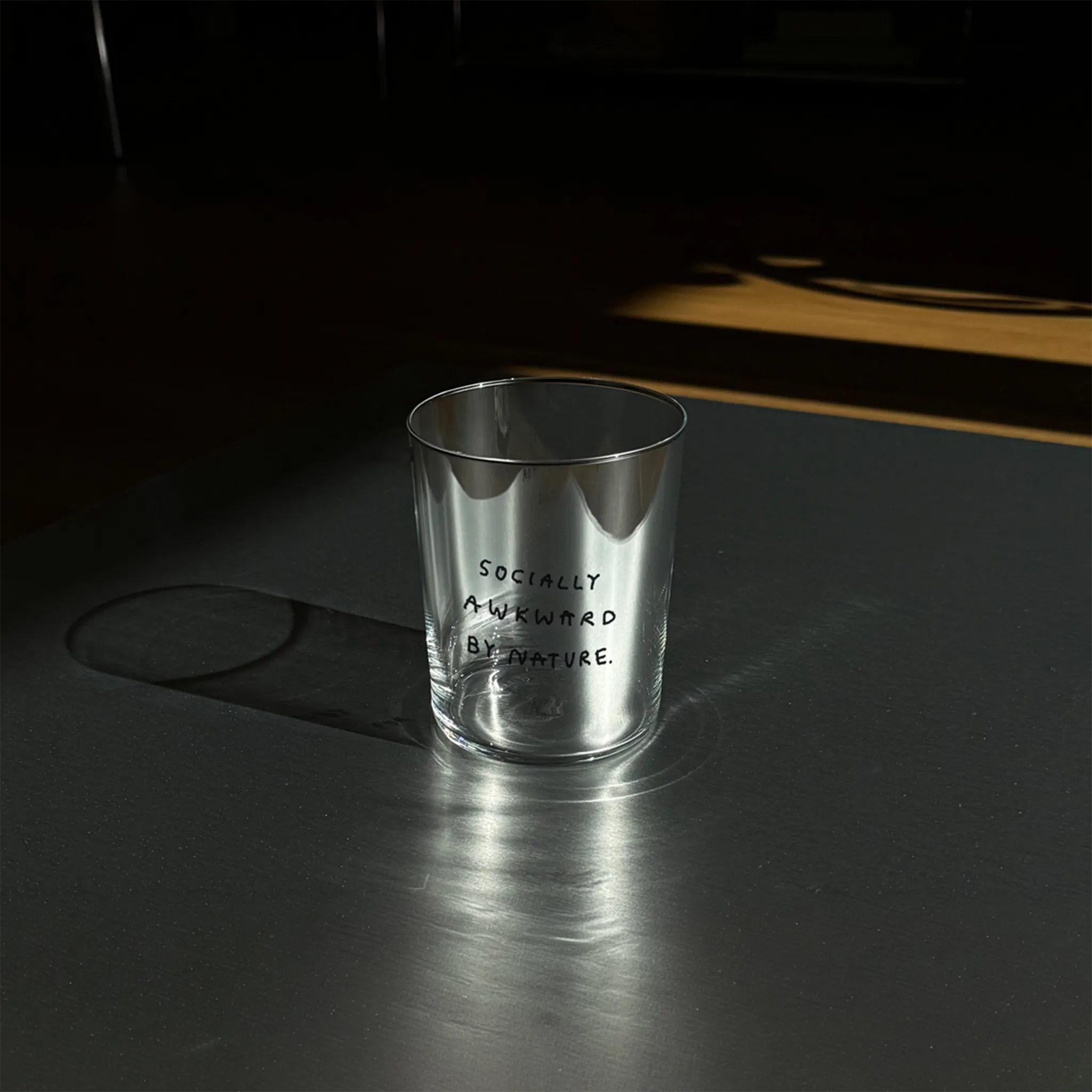 Großes Designer-Trinkglas mit sehr dünnen Glaswänden und einer schwarzen, einfarbigen Illustration auf der Vorderseite. Das Motiv auf dem Glas ist eine Handschrift in Schwarz in der geschrieben ist SOCIALLY AWKWARD BY NATURE. Das Glas steht auf einer Metallplatte.