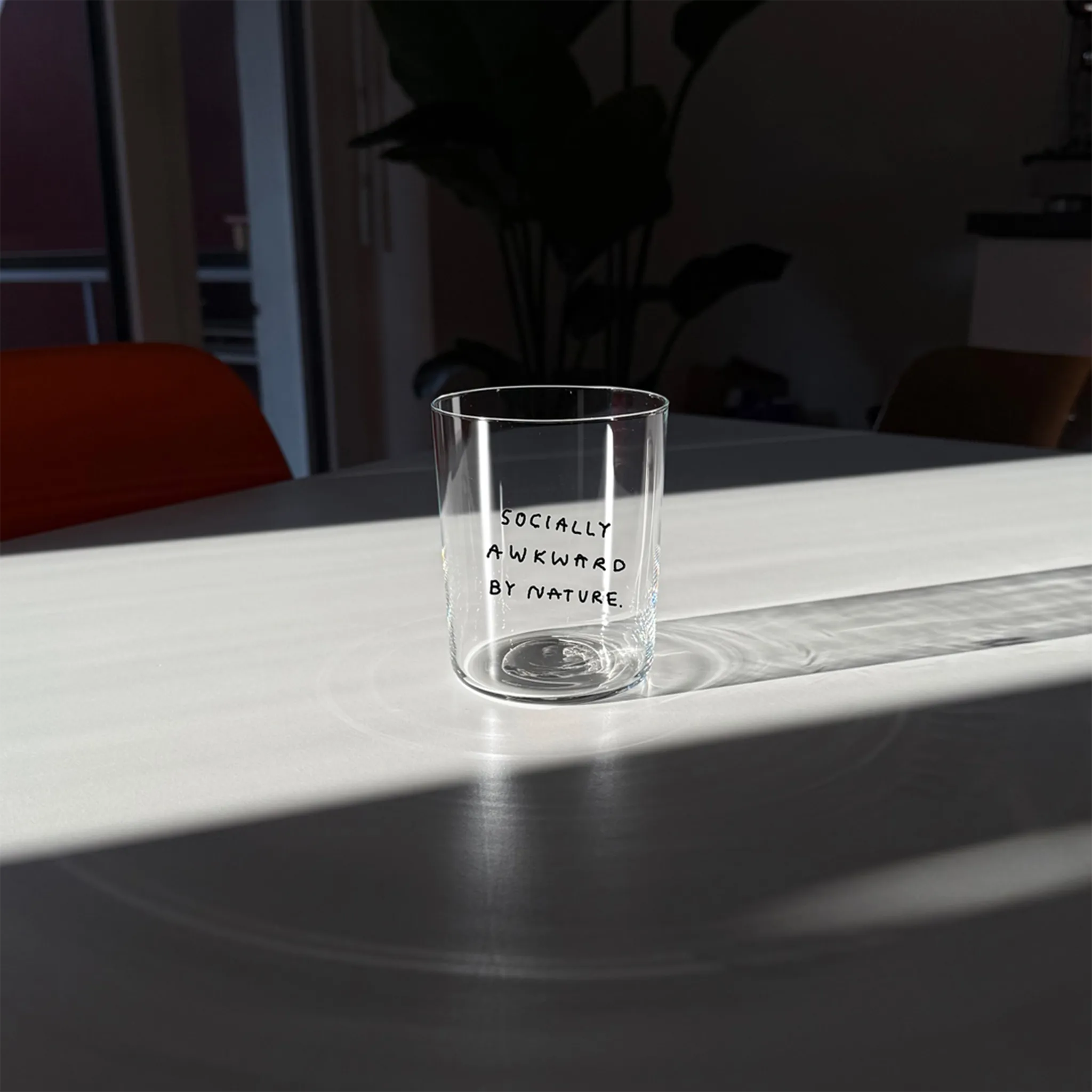 Großes Designer-Trinkglas mit sehr dünnen Glaswänden und einer schwarzen, einfarbigen Illustration auf der Vorderseite. Das Motiv auf dem Glas ist eine Handschrift in Schwarz in der geschrieben ist SOCIALLY AWKWARD BY NATURE. Das Glas steht auf einem weißen Tisch im Licht, dahinter eine große Pflanze und ein orangener Stuhl.