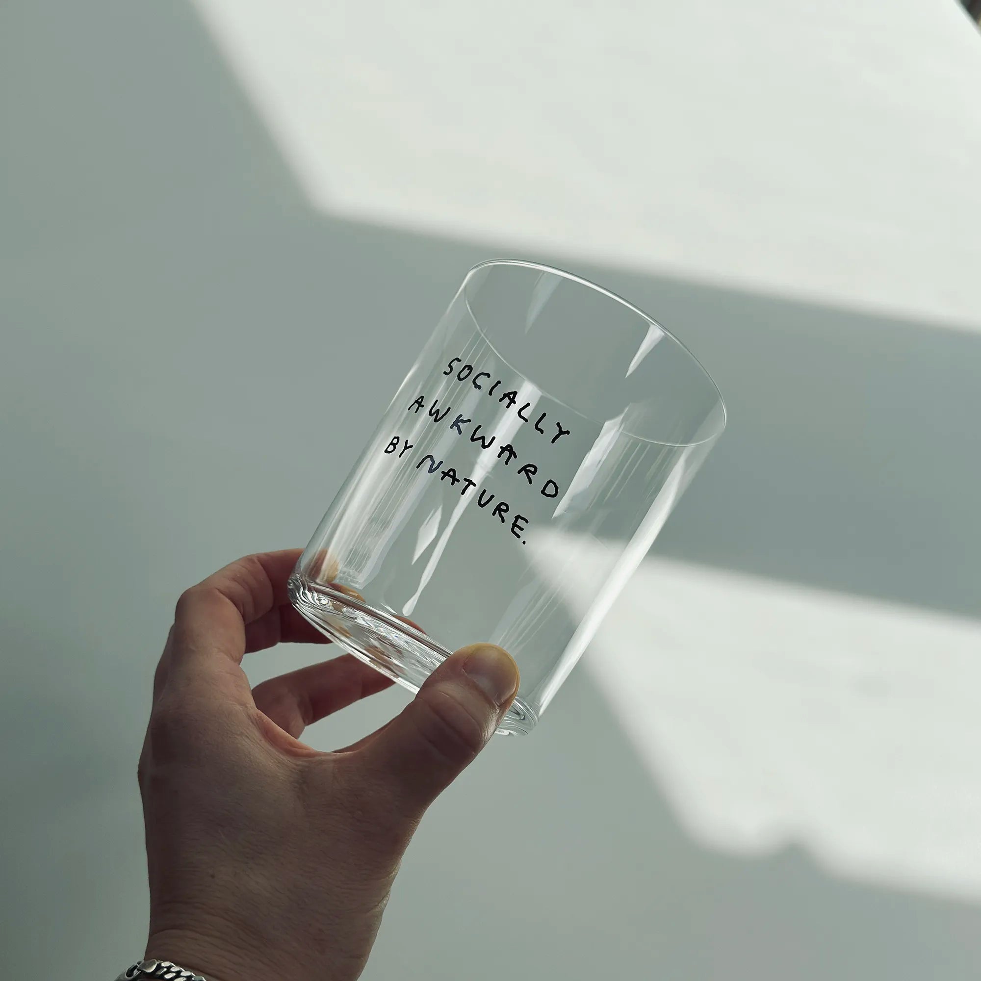 Großes Designer-Trinkglas mit sehr dünnen Glaswänden und einer schwarzen, einfarbigen Illustration auf der Vorderseite. Das Motiv auf dem Glas ist eine Handschrift in Schwarz in der geschrieben ist SOCIALLY AWKWARD BY NATURE. 

Das Glas wird über einem weißen Tisch gehalten, man sieht die Hand die sie vorsichtig festhält. 