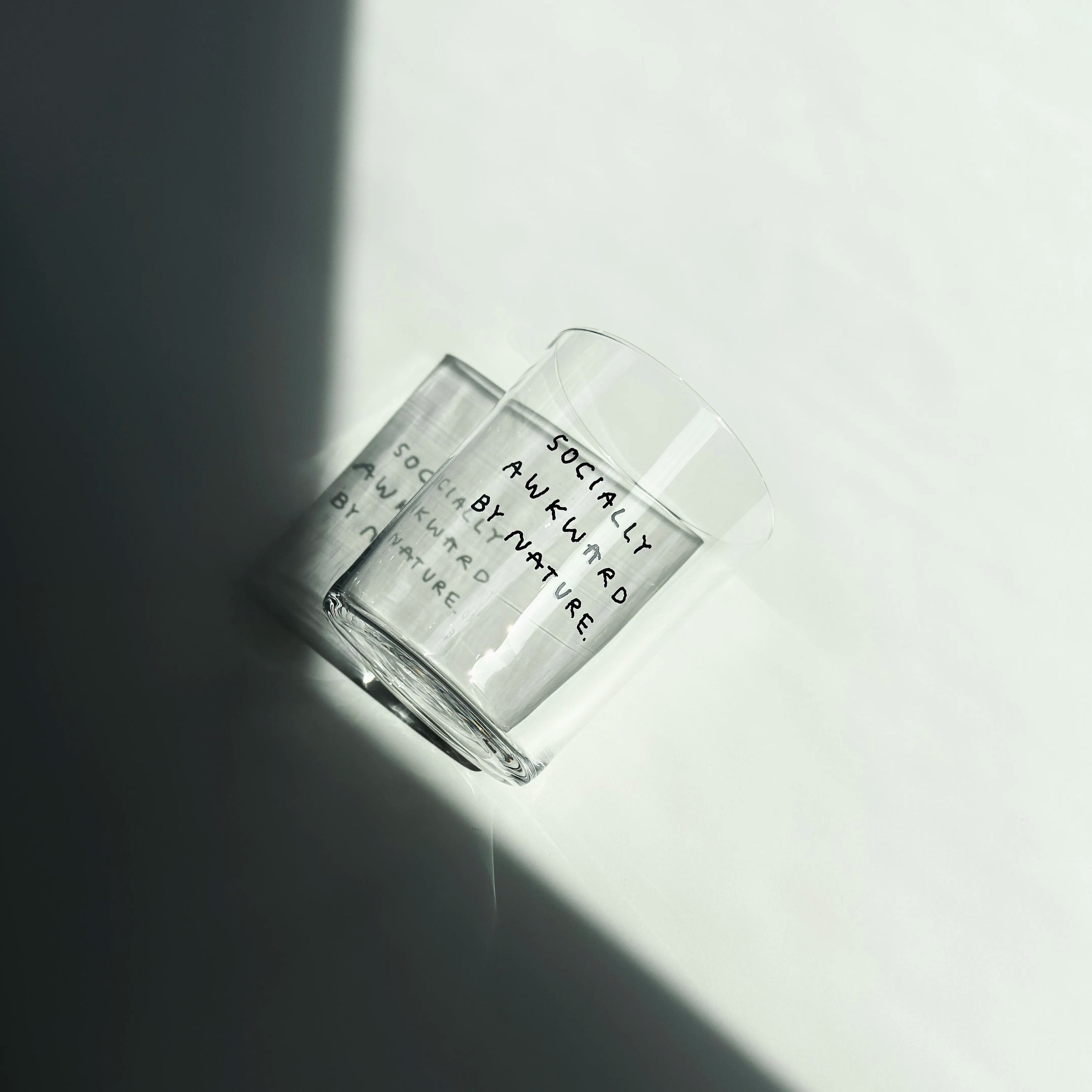 Großes Designer-Trinkglas mit sehr dünnen Glaswänden und einer schwarzen, einfarbigen Illustration auf der Vorderseite. Das Motiv auf dem Glas ist eine Handschrift in Schwarz in der geschrieben ist SOCIALLY AWKWARD BY NATURE. Das Glas steht auf einem weißen Tisch im Licht.