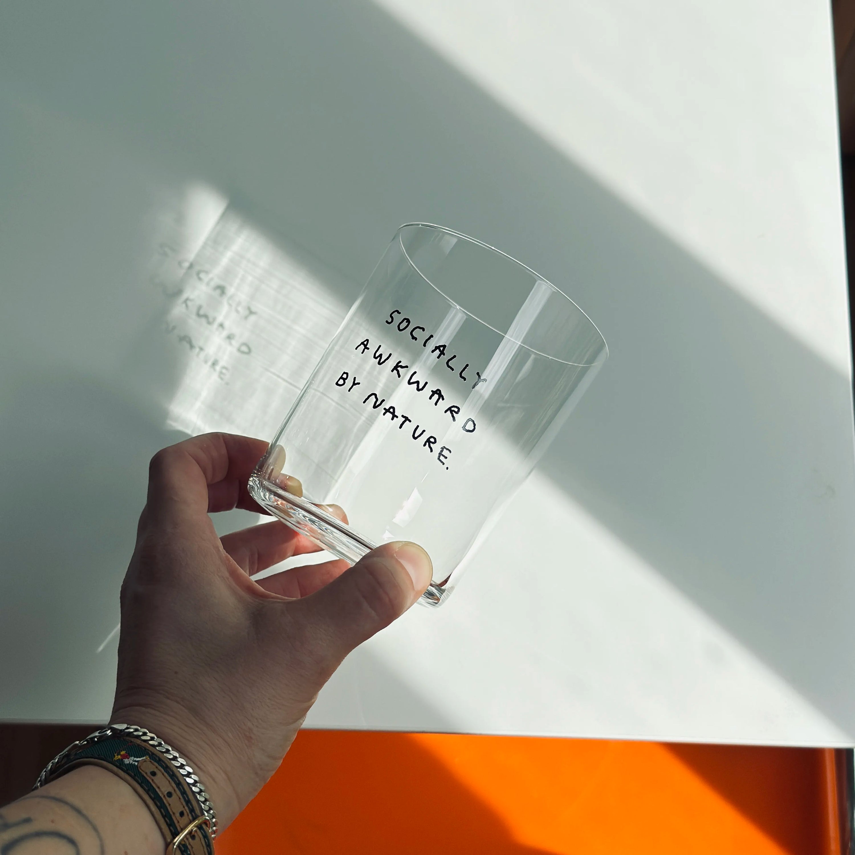 Großes Designer-Trinkglas mit sehr dünnen Glaswänden und einer schwarzen, einfarbigen Illustration auf der Vorderseite. Das Motiv auf dem Glas ist eine Handschrift in Schwarz in der geschrieben ist SOCIALLY AWKWARD BY NATURE. 

Das Glas wird über einem weißen Tisch gehalten, man sieht die Hand die sie vorsichtig festhält. 