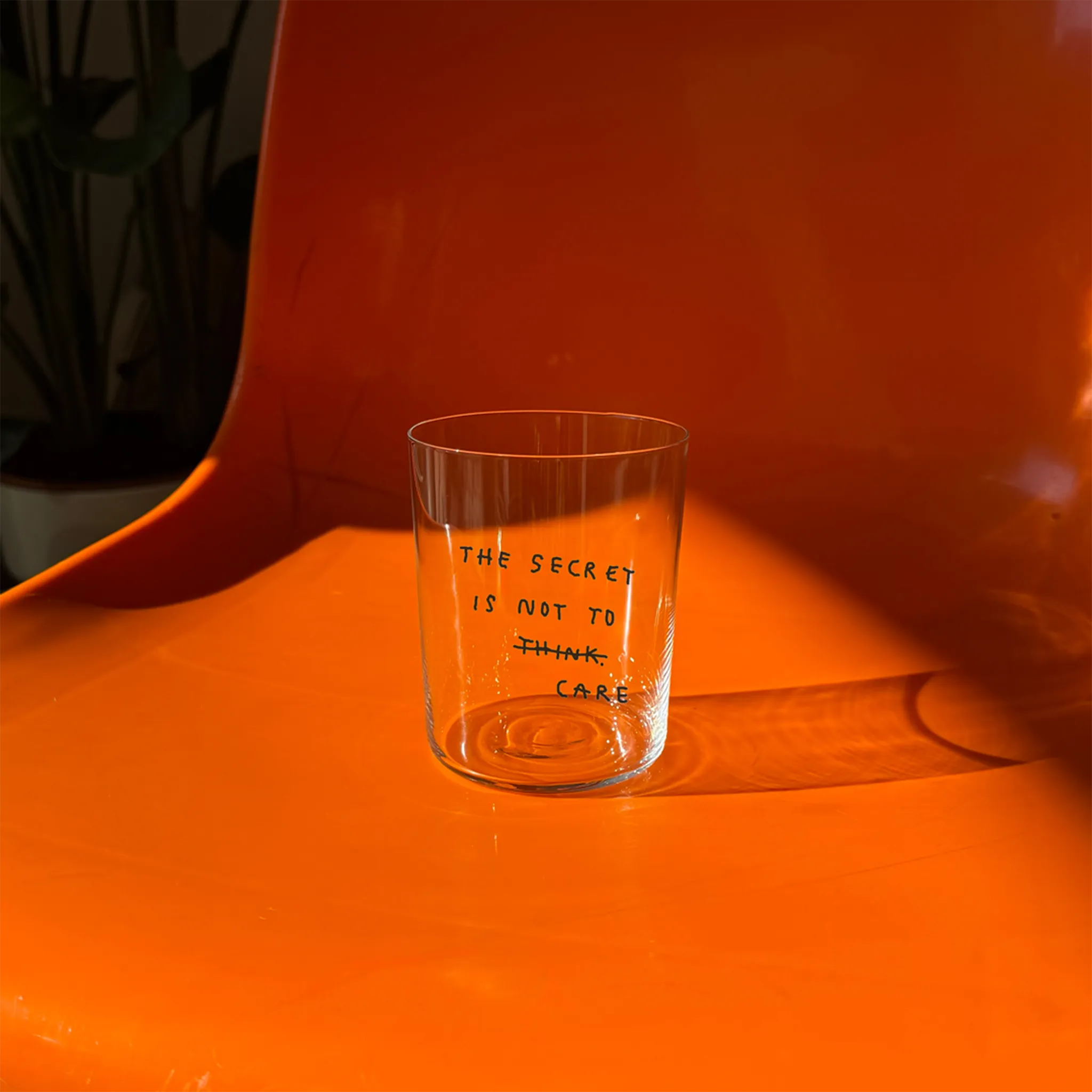 Großes Designer-Trinkglas mit sehr dünnen Glaswänden und einer schwarzen, einfarbigen Illustration auf der Vorderseite. Das Motiv auf dem Glas ist eine Handschrift in Schwarz in der geschrieben ist The secret is not to think wobei das think durchgestrichen ist und drunter das Wort CARE steht. Das Glas steht auf einem orangenen Stuhl.