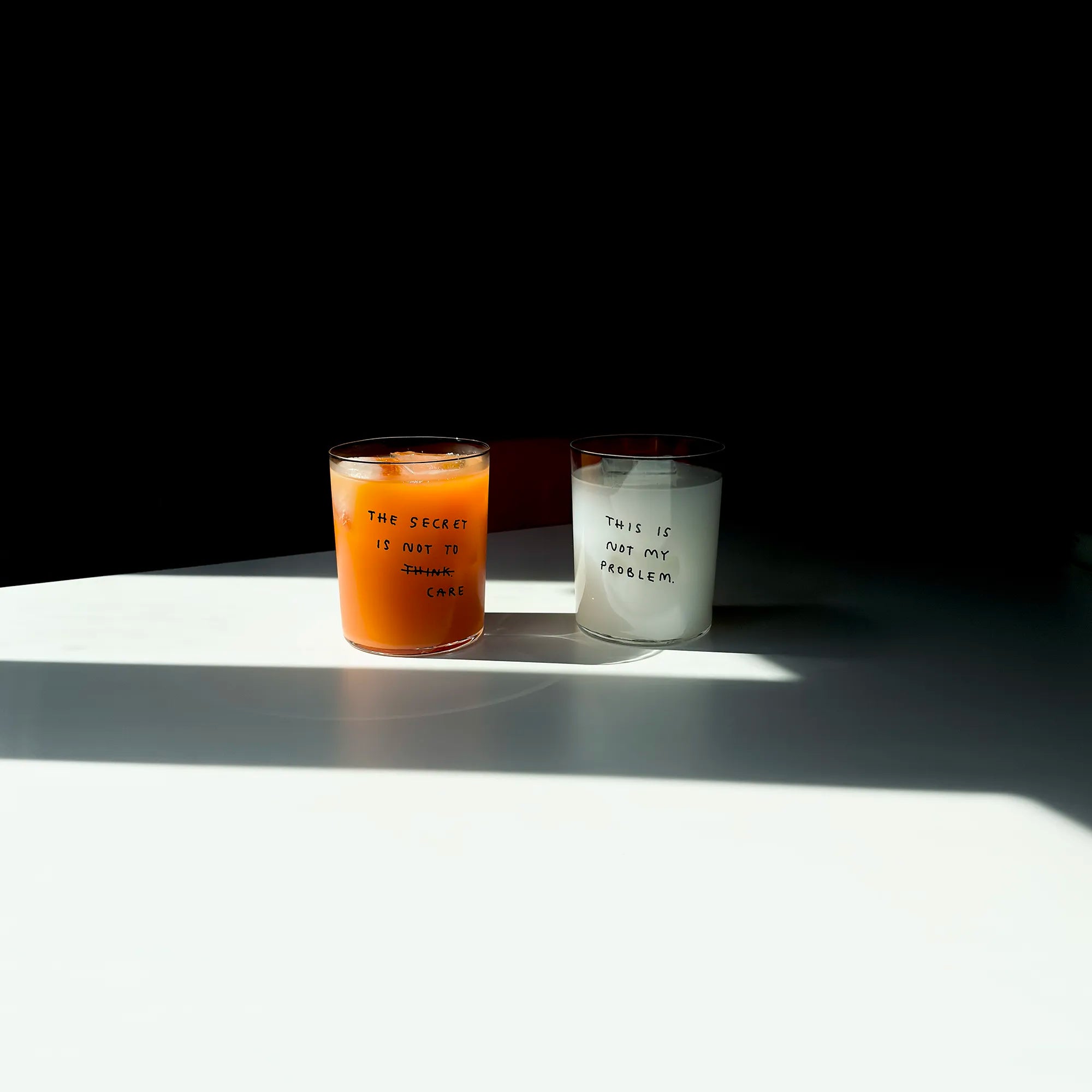 Großes Designer-Trinkglas mit sehr dünnen Glaswänden und einer schwarzen, einfarbigen Illustration auf der Vorderseite. Das Motiv auf dem Glas ist eine Handschrift in Schwarz in der geschrieben ist The secret is not to think wobei das think durchgestrichen ist und drunter das Wort CARE steht. Das Glas liegt auf einer Metallplatte und wirft einen schönen Schatten.

Das Glas ist mit einer orangenen Flüssigkeit und Eiswürfeln befüllt, daneben steht ein Glas mit der Aufschrift THIS IS NOT MY PROBLEM.