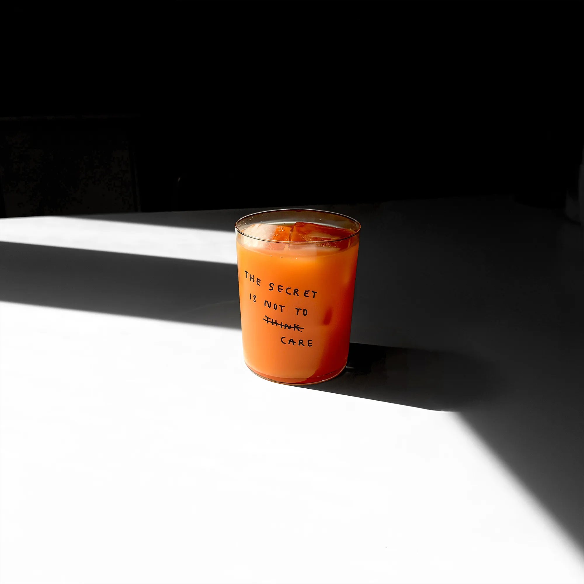 Großes Designer-Trinkglas mit sehr dünnen Glaswänden und einer schwarzen, einfarbigen Illustration auf der Vorderseite. Das Motiv auf dem Glas ist eine Handschrift in Schwarz in der geschrieben ist The secret is not to think wobei das think durchgestrichen ist und drunter das Wort CARE steht. Das Glas liegt auf einer Metallplatte und wirft einen schönen Schatten.

Das Glas ist mit einer orangenen Flüssigkeit und Eiswürfeln befüllt.