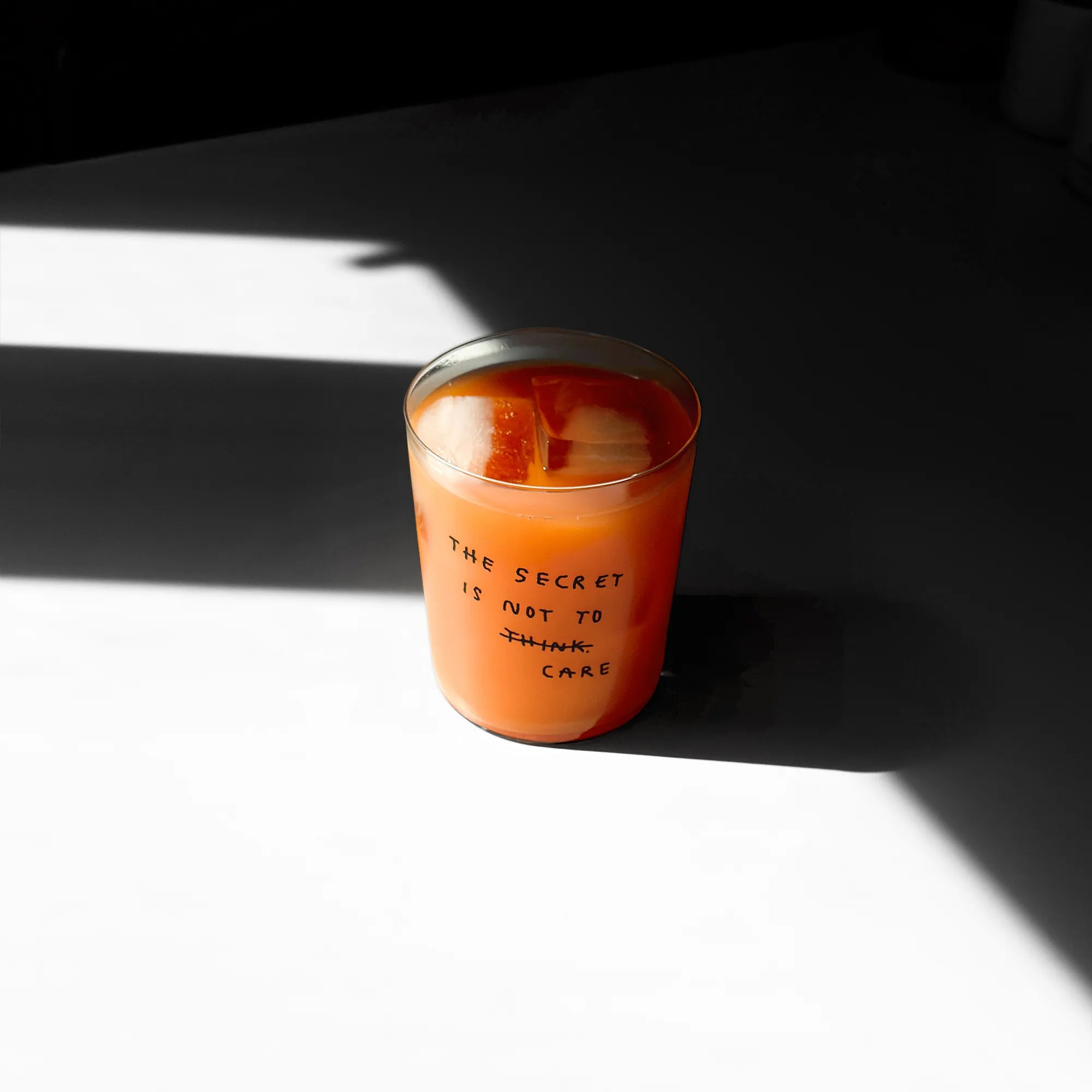 Großes Designer-Trinkglas mit sehr dünnen Glaswänden und einer schwarzen, einfarbigen Illustration auf der Vorderseite. Das Motiv auf dem Glas ist eine Handschrift in Schwarz in der geschrieben ist The secret is not to think wobei das think durchgestrichen ist und drunter das Wort CARE steht. Das Glas liegt auf einer Metallplatte und wirft einen schönen Schatten.

Das Glas ist mit einer orangenen Flüssigkeit und Eiswürfeln befüllt.
