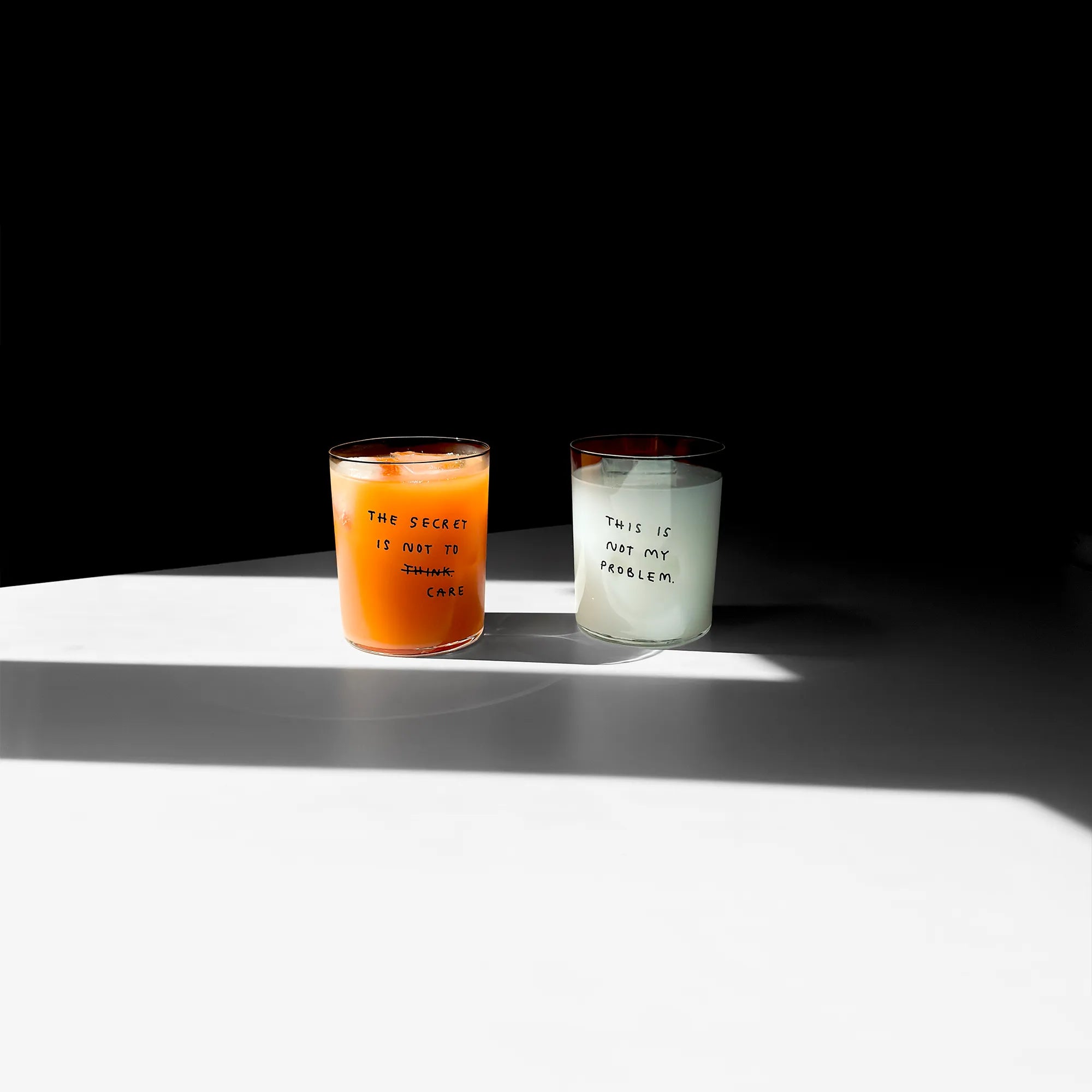 Großes Designer-Trinkglas mit sehr dünnen Glaswänden und einer schwarzen, einfarbigen Illustration auf der Vorderseite. Das Motiv auf dem Glas ist eine Handschrift in Schwarz in der geschrieben ist The secret is not to think wobei das think durchgestrichen ist und drunter das Wort CARE steht. Das Glas liegt auf einer Metallplatte und wirft einen schönen Schatten.

Das Glas ist mit einer orangenen Flüssigkeit und Eiswürfeln befüllt, daneben steht ein Glas mit der Aufschrift THIS IS NOT MY PROBLEM.