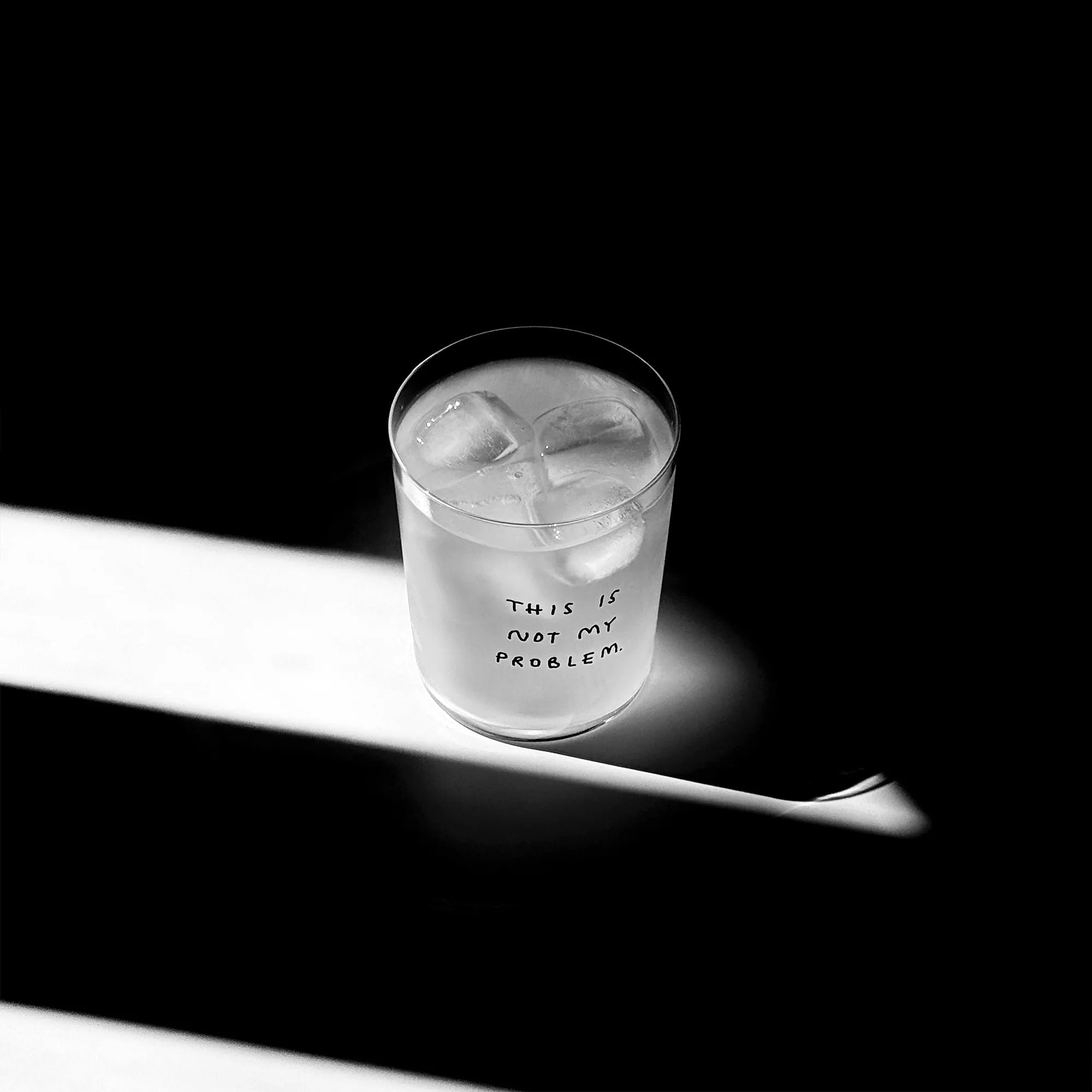 Großes Designer-Trinkglas mit sehr dünnen Glaswänden und einer schwarzen, einfarbigen Illustration auf der Vorderseite mit dem Motiv „This is not my problem“. Illustration von Künstlerin, Illustratorin und Co-Gründerin Johanna Schwarzer.

Das Glas steht auf einem weißen Tisch, das Foto ist in einer Nahaufnahme, das Glas ist mit einer transparenten Flüssigkeit und Eiswürfeln befüllt. Im Hintergrund viel Schatten. Das Glas steht mitten in einem Lichtwurf.