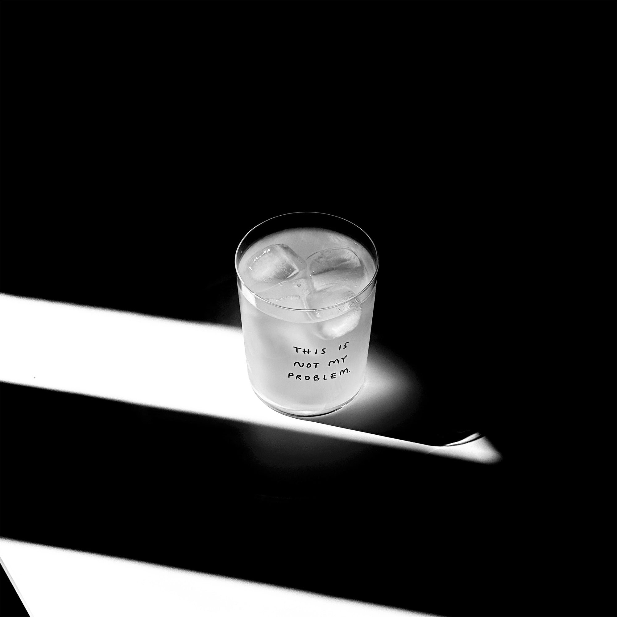 Großes Designer-Trinkglas mit sehr dünnen Glaswänden und einer schwarzen, einfarbigen Illustration auf der Vorderseite mit dem Motiv „This is not my problem“. Illustration von Künstlerin, Illustratorin und Co-Gründerin Johanna Schwarzer.

Das Glas steht auf einem weißen Tisch, das Foto ist in einer Nahaufnahme, das Glas ist mit einer transparenten Flüssigkeit und Eiswürfeln befüllt. Im Hintergrund viel Schatten. Das Glas steht mitten in einem Lichtwurf.