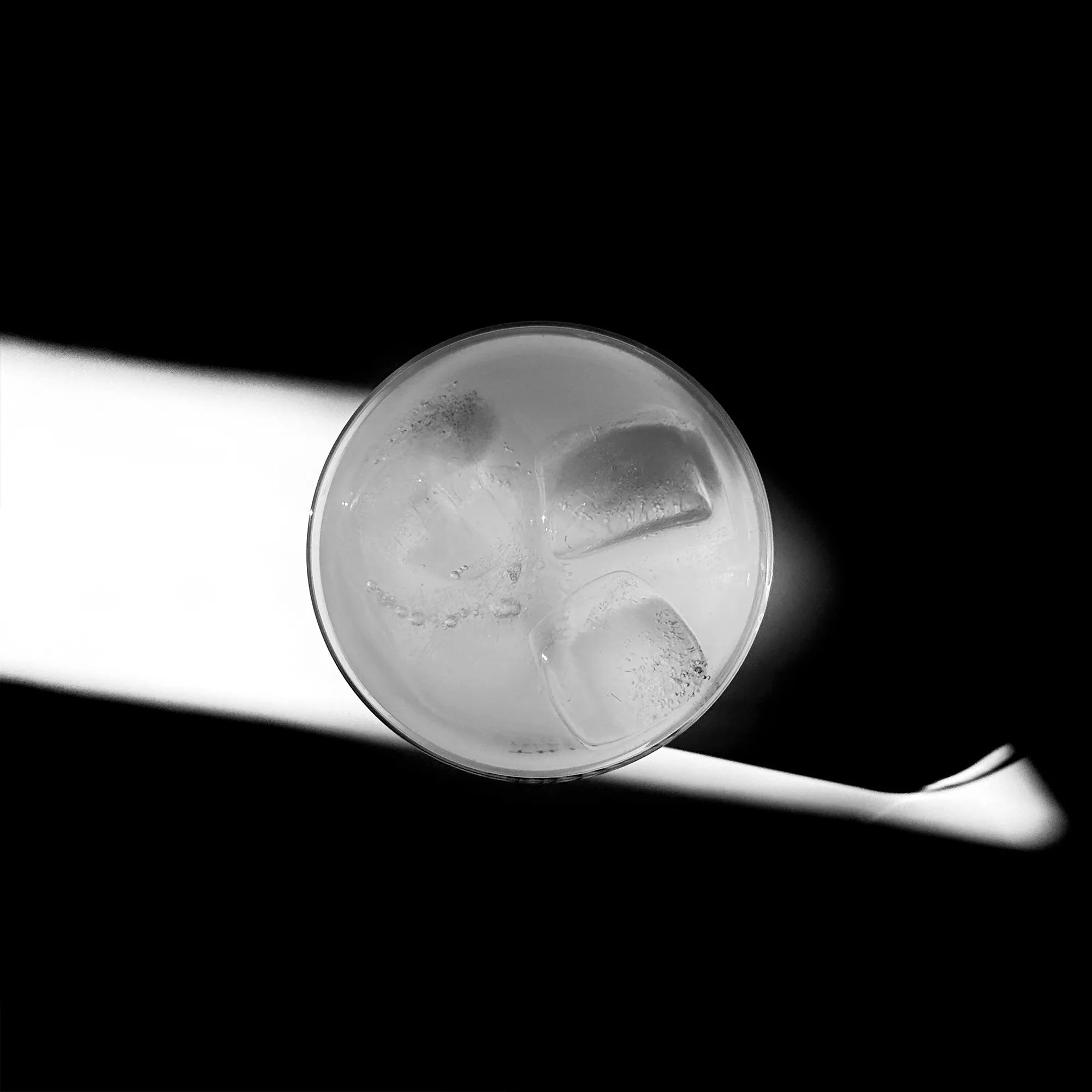 Fotoaufnahme eines Glasses von oben. Das Glas ist mit einer transparenten Flüssigkeit und Eiswürfeln befüllt. Das Glas steht mitten in einem Lichtwurf, drum herum nur Schatten.