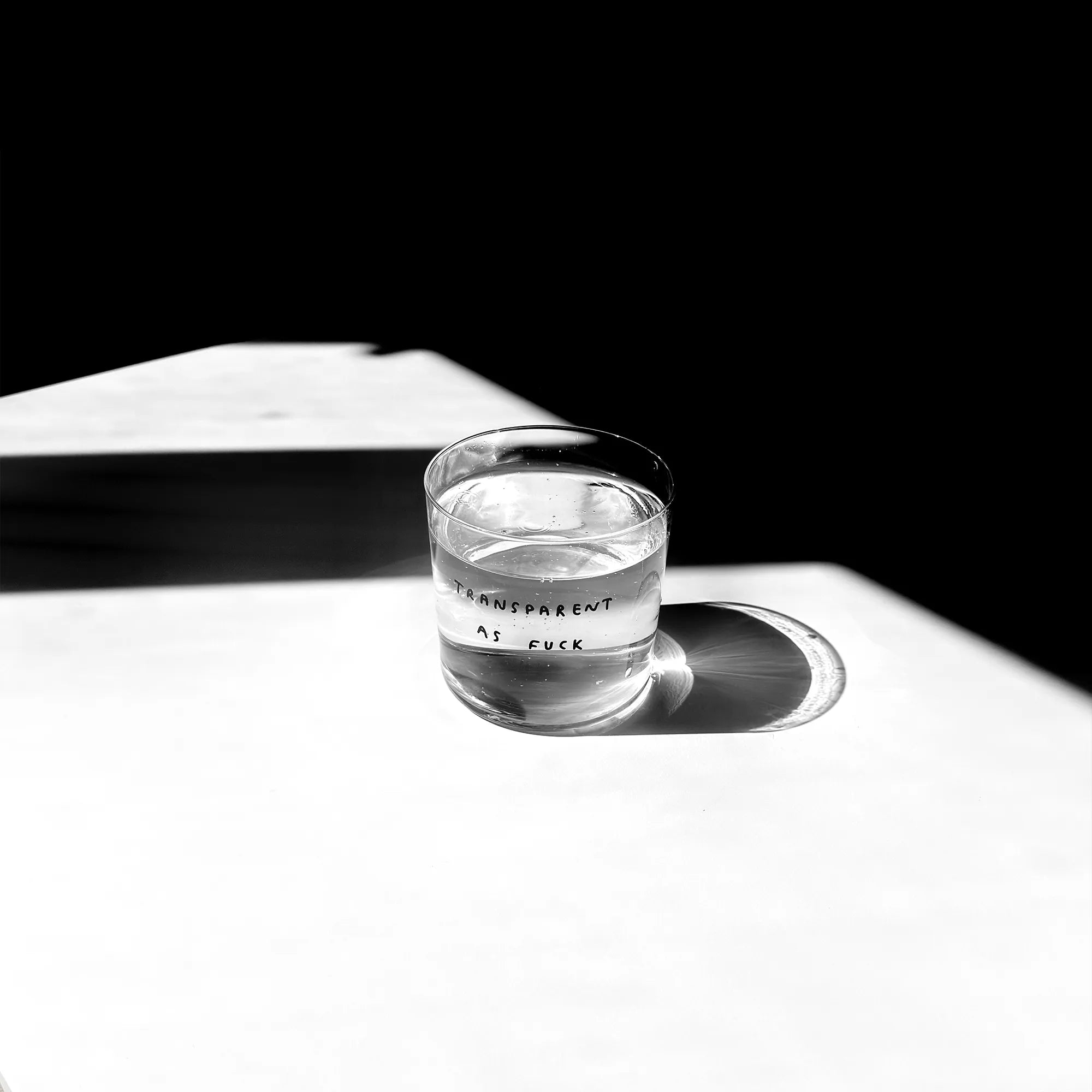 Großes Designer-Trinkglas mit sehr dünnen Glaswänden und einer schwarzen, einfarbigen Illustration auf der Vorderseite mit dem Motiv „Transparent as fuck. Illustration von Künstlerin, Illustratorin und Co-Gründerin Johanna Schwarzer.

Das Glas steht auf einem weißen Tisch, das Foto ist in einer Nahaufnahme, das Glas ist mit einer transaprenten Flüssigkeit befüllt. Im Hintergrund viel Schatten.