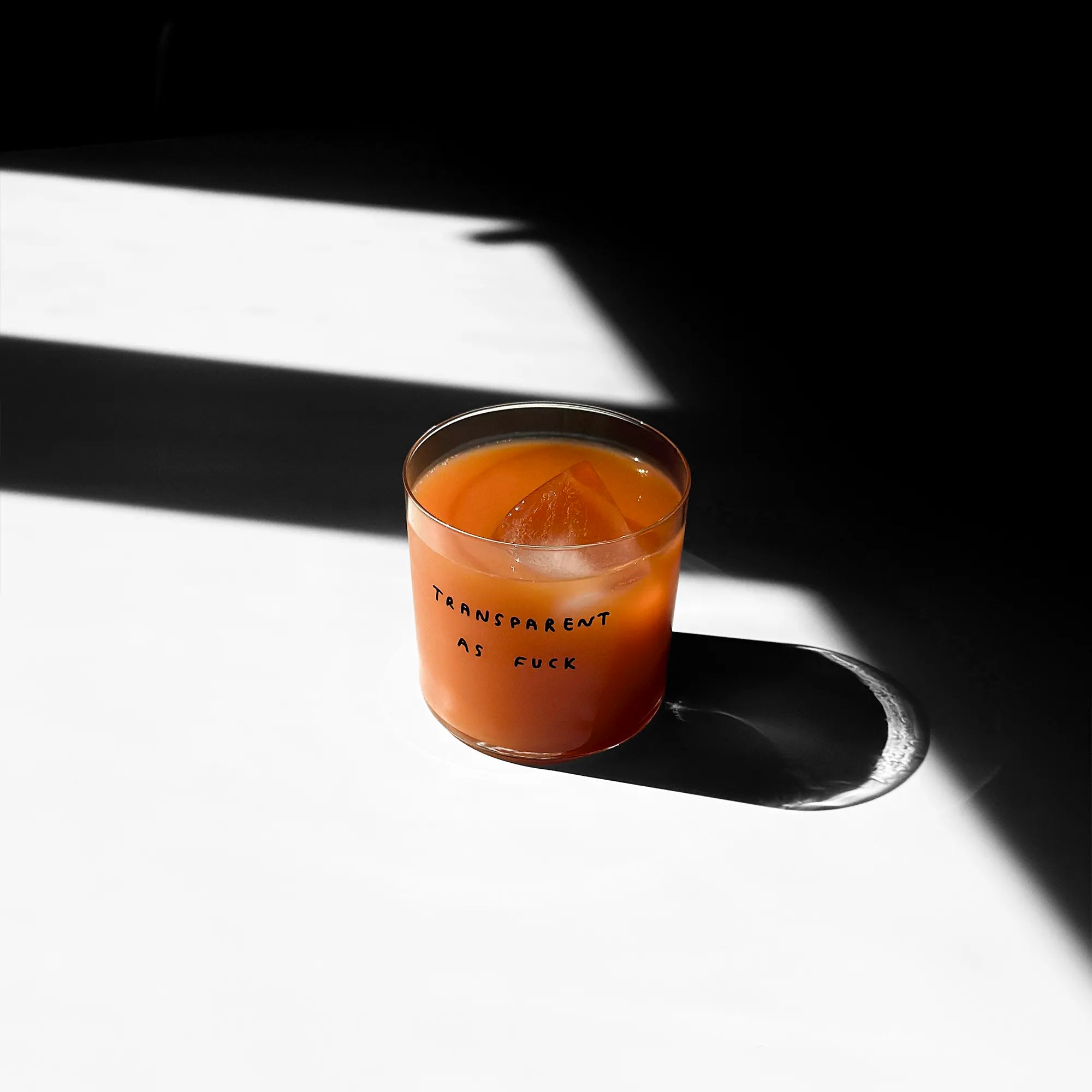 Medium Designer-Trinkglas mit sehr dünnen Glaswänden und einer schwarzen, einfarbigen Illustration auf der Vorderseite. Das Motiv zeigt eine Typografie in der geschrieben ist TRANSPARENT AS FUCK.

Das Glas steht auf einem weißen Tisch und ist mit einer Orangenen Flüssigkeit gefüllt. Ein Eiswürfel ist im Glas, das Glas wirft einen großen Schatten. 