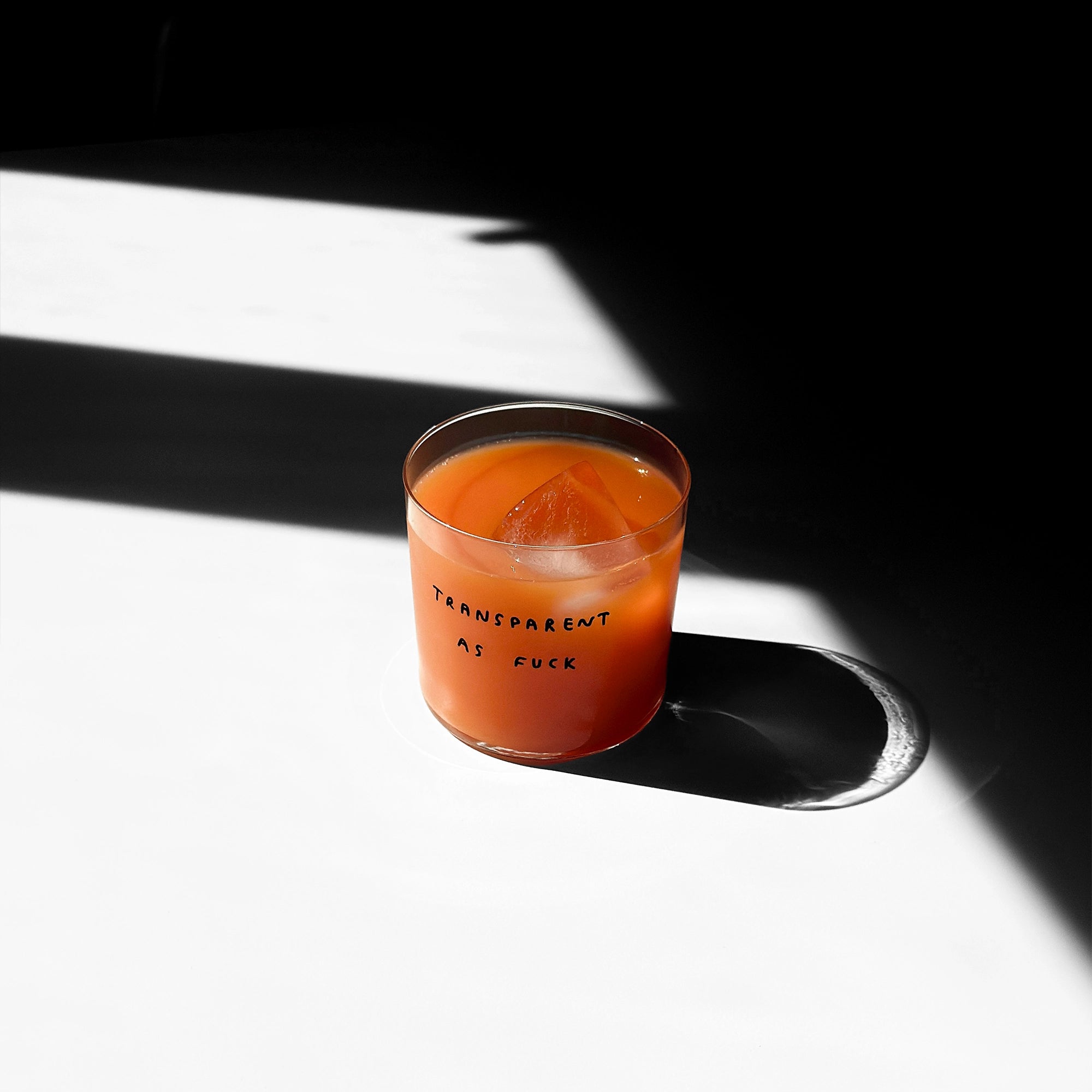 Medium Designer-Trinkglas mit sehr dünnen Glaswänden und einer schwarzen, einfarbigen Illustration auf der Vorderseite. Das Motiv zeigt eine Typografie in der geschrieben ist TRANSPARENT AS FUCK.

Das Glas steht auf einem weißen Tisch und ist mit einer Orangenen Flüssigkeit gefüllt. Ein Eiswürfel ist im Glas, das Glas wirft einen großen Schatten. 