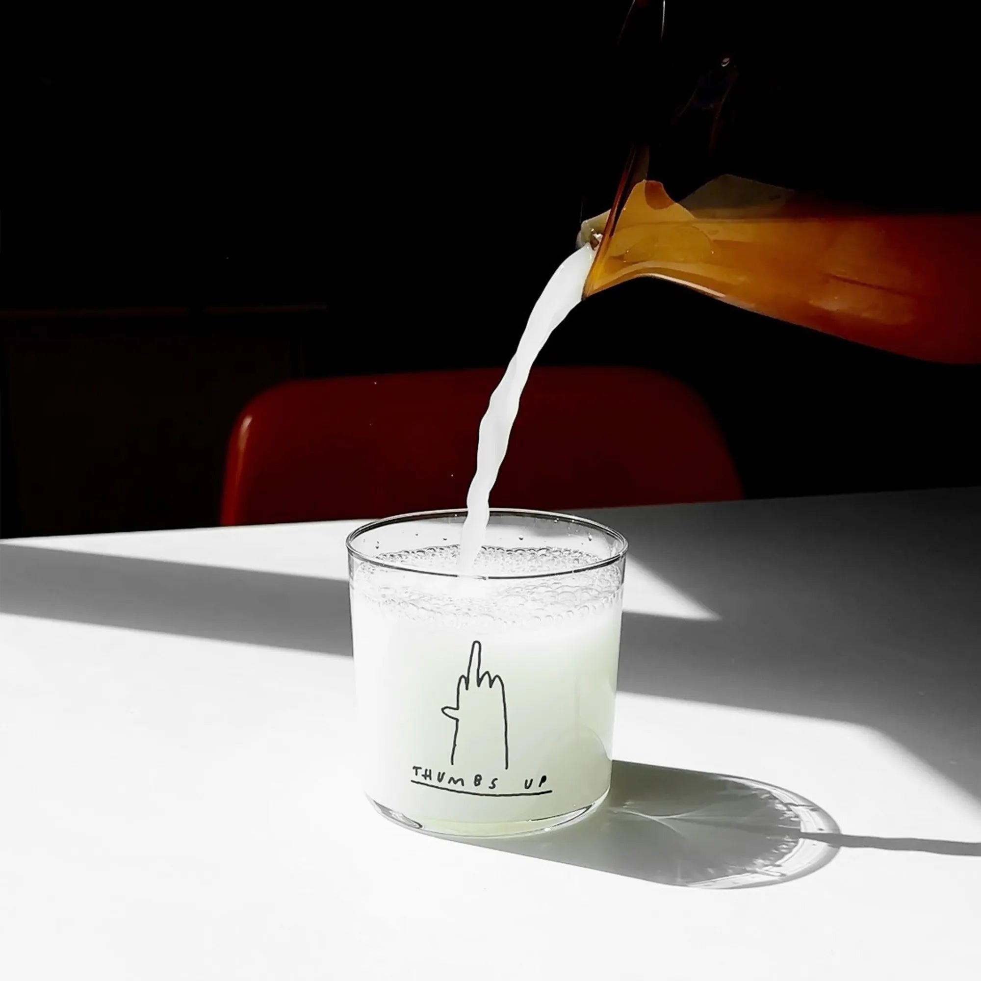 Medium Designer-Trinkglas mit sehr dünnen Glaswänden und einer schwarzen, einfarbigen Illustration auf der Vorderseite. Das Motiv zeigt eine Typografie in der geschrieben THUMBS UP und einer Illustration einer Hand die den Mittelfinger zeigt.

Das Glas ist mit einer weißen Flüssigkeit befüllt, steht auf einem Weißen Tisch in einem Lichtkegel, dahinter ist ein Stuhl ganz dezent erkennbar. 
