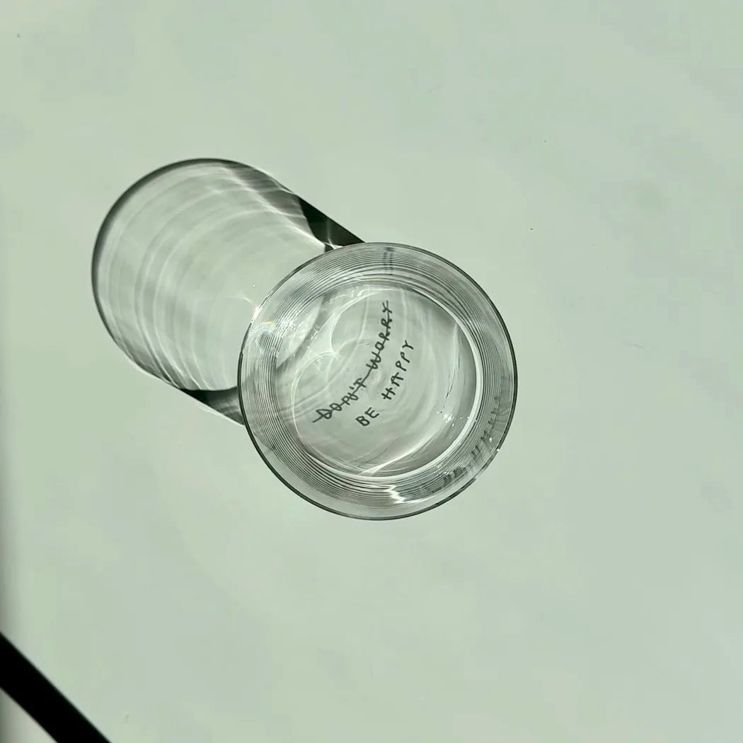 Transparentes Medium Designer Glas mit schwarzem Schriftzug "DONT WORRY BE HAPPY" wobei DONT WORRY durchgestrichen ist und lediglich die Aufforderung BE HAPPY im Vordergrund steht, die Gläser haben dünnen Wänden und Boden. 
 
Das Glas ist mit unbefüllt, das Foto wird von oben geschossen. Das Motiv ist lediglich als Schattenwurf im Glas sichtbar. 