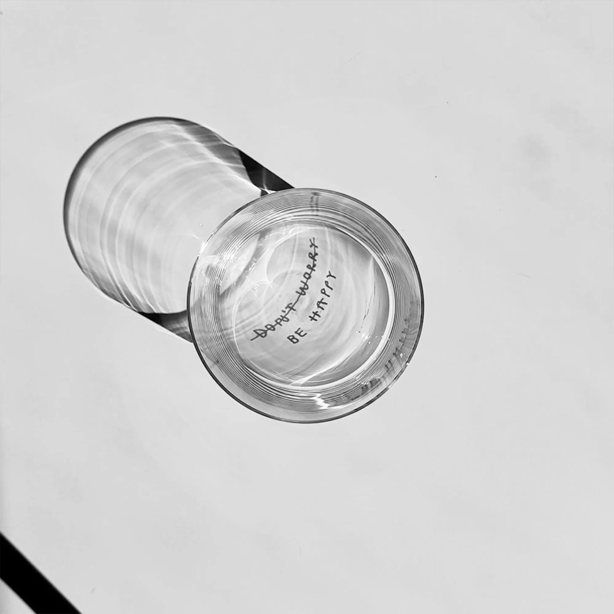 Transparentes Medium Designer Glas mit schwarzem Schriftzug "DONT WORRY BE HAPPY" wobei DONT WORRY durchgestrichen ist und lediglich die Aufforderung BE HAPPY im Vordergrund steht, die Gläser haben dünnen Wänden und Boden. 
 
Das Glas ist mit unbefüllt, das Foto wird von oben geschossen. Das Motiv ist lediglich als Schattenwurf im Glas sichtbar. 