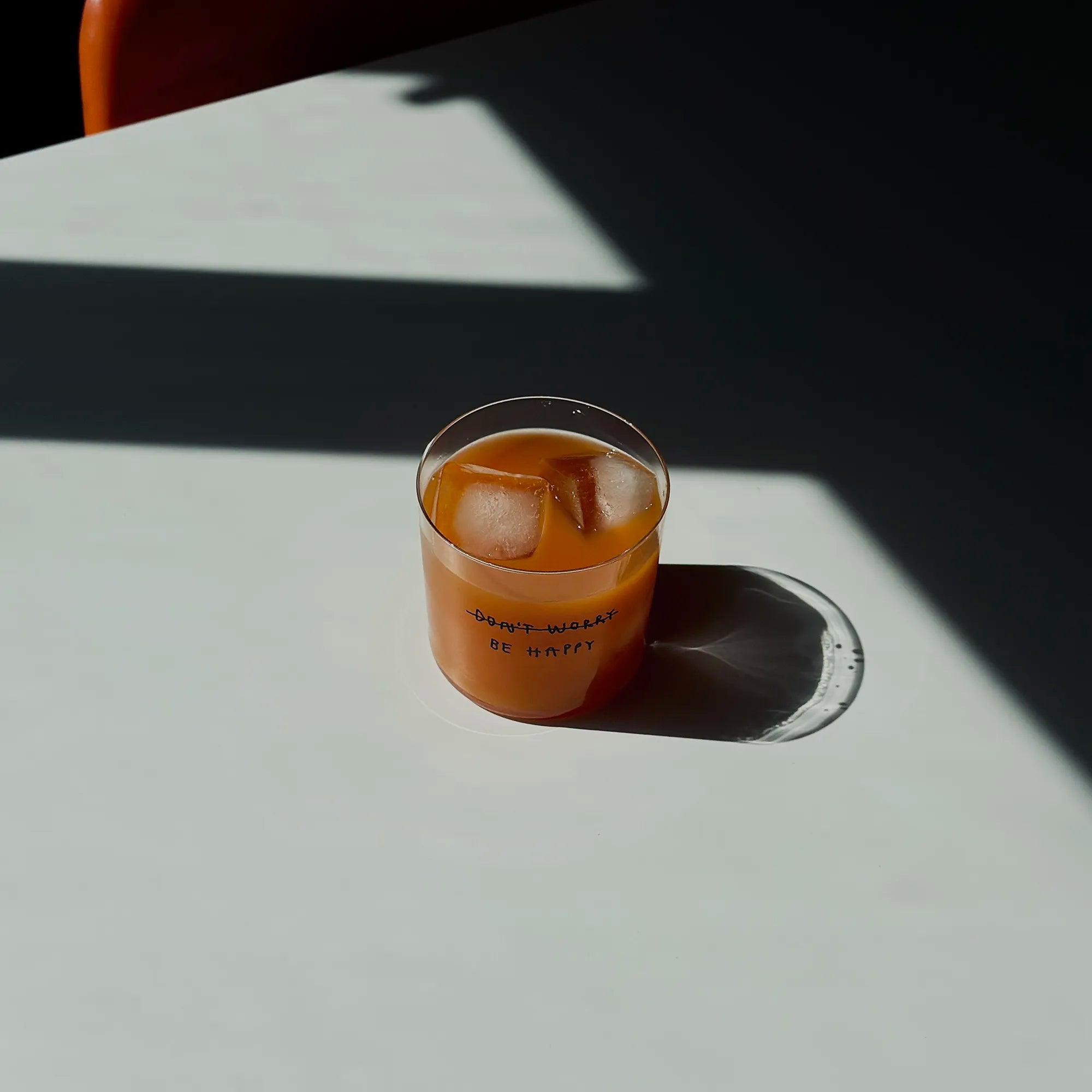 Transparentes Medium Designer Glas mit schwarzem Schriftzug "DONT WORRY BE HAPPY" wobei DONT WORRY durchgestrichen ist und lediglich die Aufforderung BE HAPPY im Vordergrund steht, die Gläser haben dünnen Wänden und Boden. 
 
Das Glas ist mit einer orangenen Flüssigkeit und Eiswürfeln befüllt und steht auf einem weißen Tisch, daneben ein orangener Stuhl. 