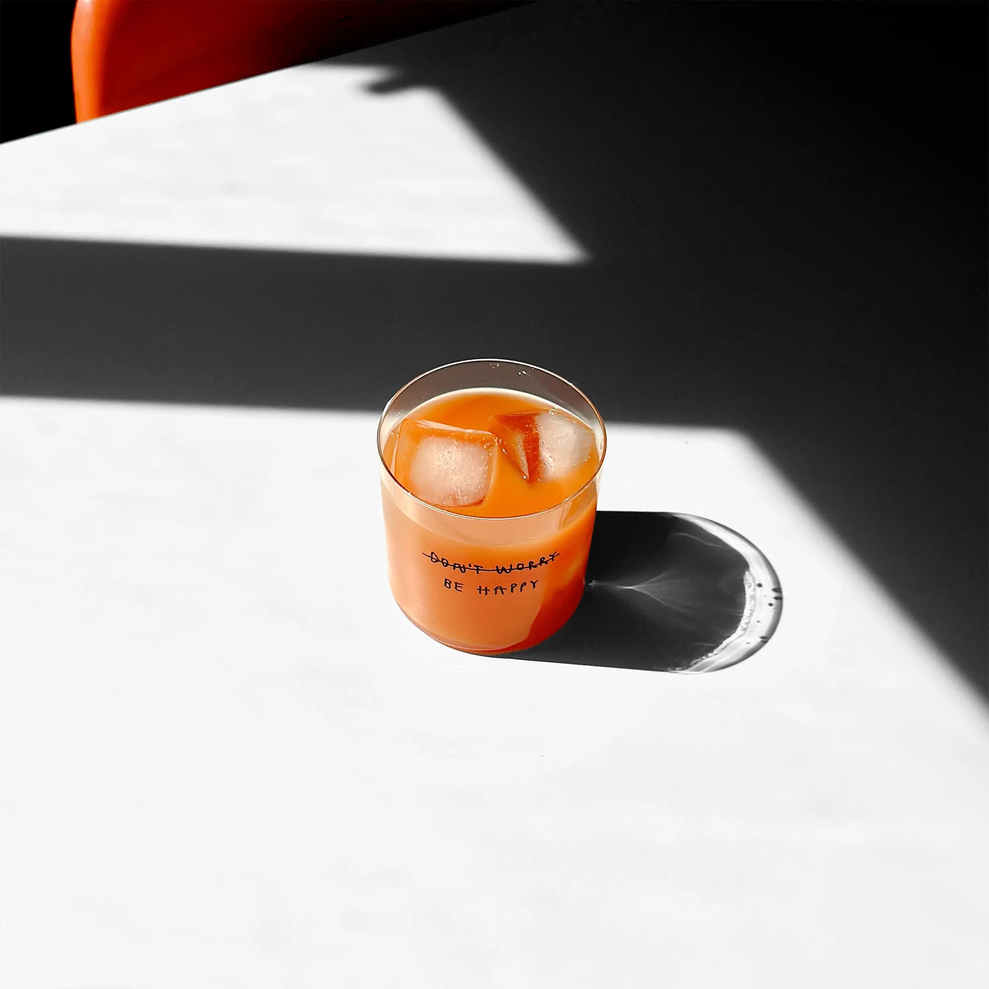 Transparentes Medium Designer Glas mit schwarzem Schriftzug "DONT WORRY BE HAPPY" wobei DONT WORRY durchgestrichen ist und lediglich die Aufforderung BE HAPPY im Vordergrund steht, die Gläser haben dünnen Wänden und Boden. 
 
Das Glas ist mit einer orangenen Flüssigkeit und Eiswürfeln befüllt und steht auf einem weißen Tisch, daneben ein orangener Stuhl. 