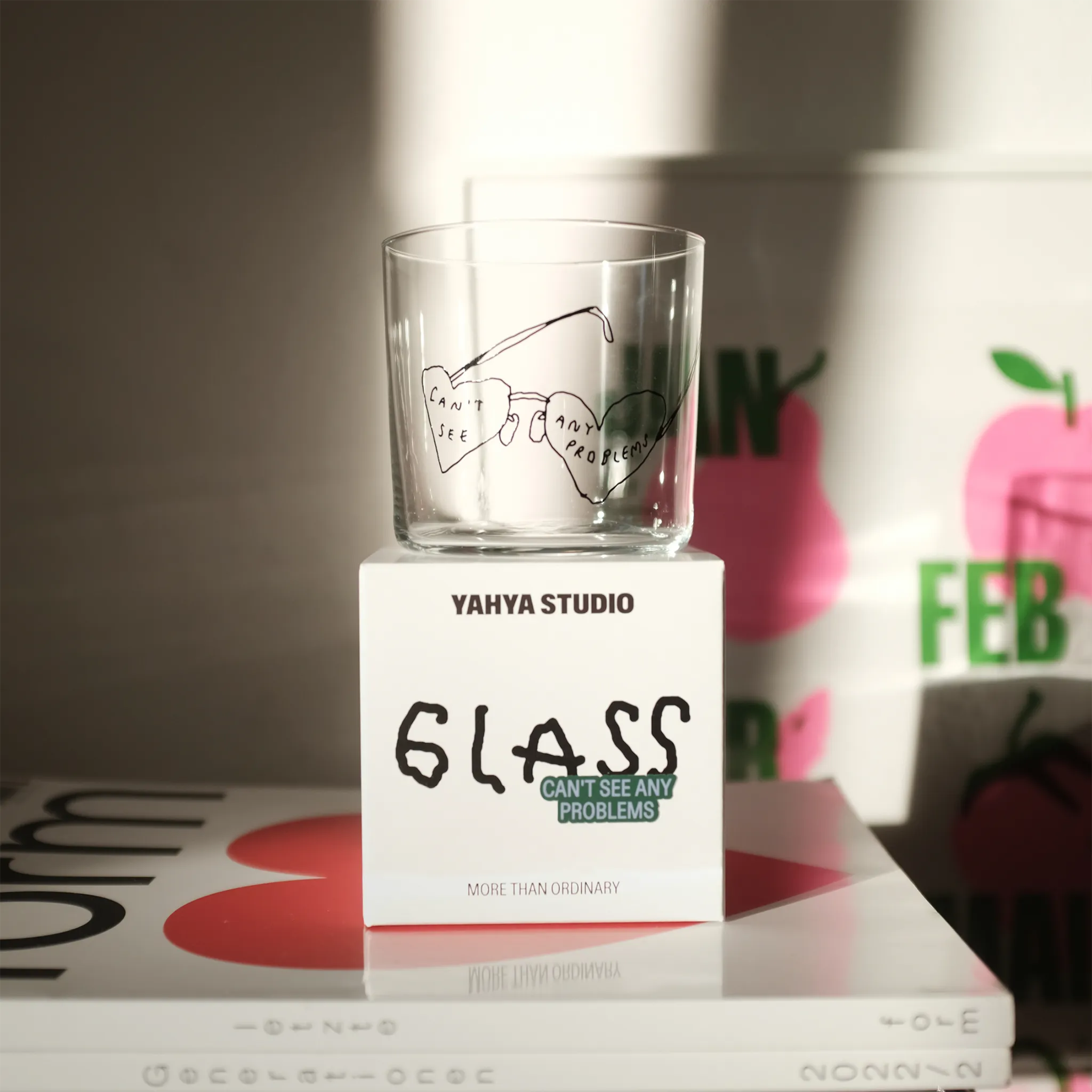 Transparentes Medium Designer Glas mit schwarzem Schriftzug "can't see any problems" mit einer Brille, die Brillengläser in herzformen hat. Das Glas steht auf seiner weißen Verpackung auf einem Buch. 