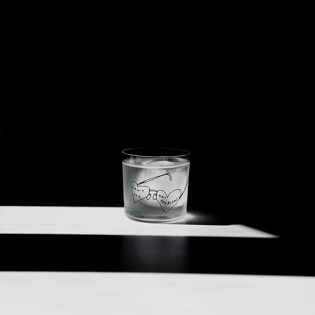 Transparentes Medium Designer Glas mit schwarzem Schriftzug "can't see any problems" mit einer Brille, die Brillengläser in herzformen hat. Das Glas steht in einem Lichtkegel, viel Schatten im Hintergrund, auf einem weißen Tisch.