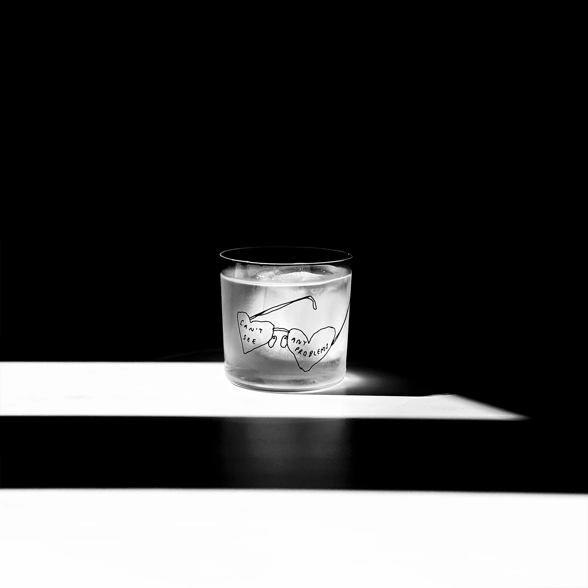 Transparentes Medium Designer Glas mit schwarzem Schriftzug "can't see any problems" mit einer Brille, die Brillengläser in herzformen hat. Das Glas steht in einem Lichtkegel, viel Schatten im Hintergrund, auf einem weißen Tisch.