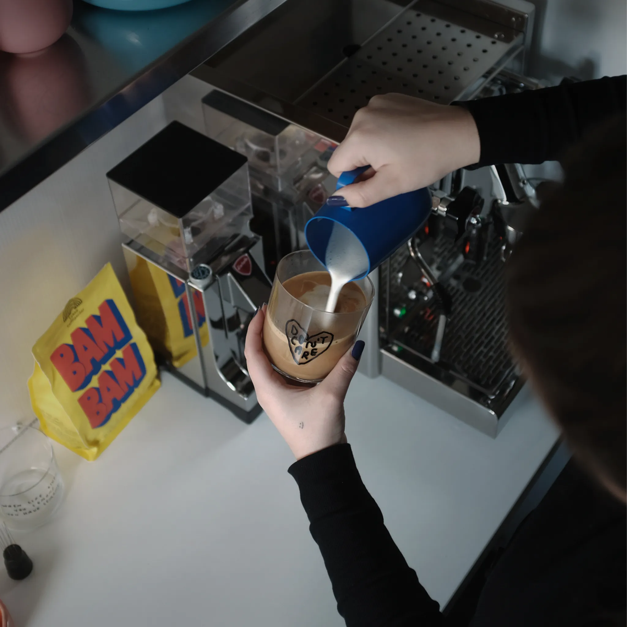 Transparentes Medium Designer Glas mit schwarzem Schriftzug "DON'T CARE" in einem Herz mit dünnen Wänden und Boden. 

Das Glas wird mit aufgeschäumter Milch befüllt und in der Hand gehalten. Im Hintergrund sieht man eine Siebträgermaschine sowie eine gelbe Packung Kaffeebohnen. 