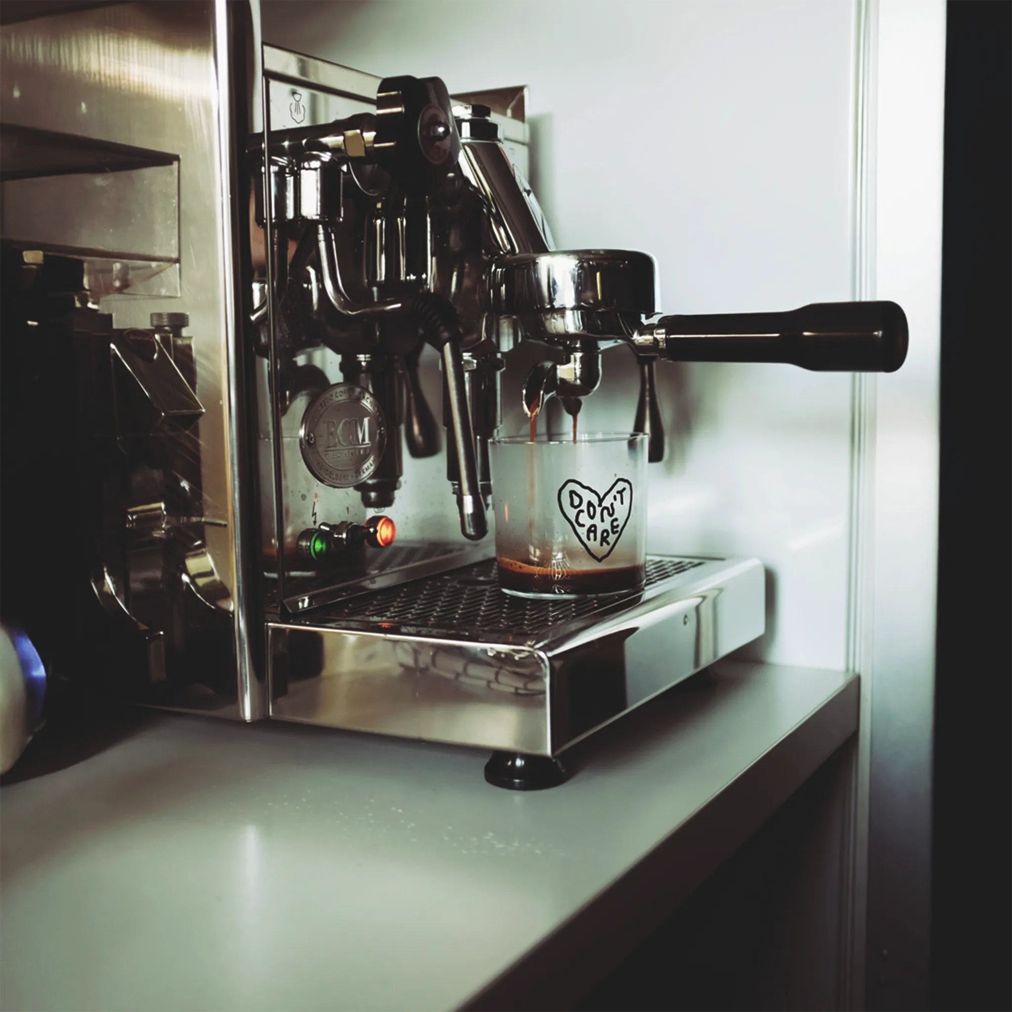 Transparentes Medium Designer Glas mit schwarzem Schriftzug "DON'T CARE" in einem Herz mit dünnen Wänden und Boden. 

Das Glas steht unter einer Siebträger-Kaffeemaschine aus der Kaffee in das glas läuft. 