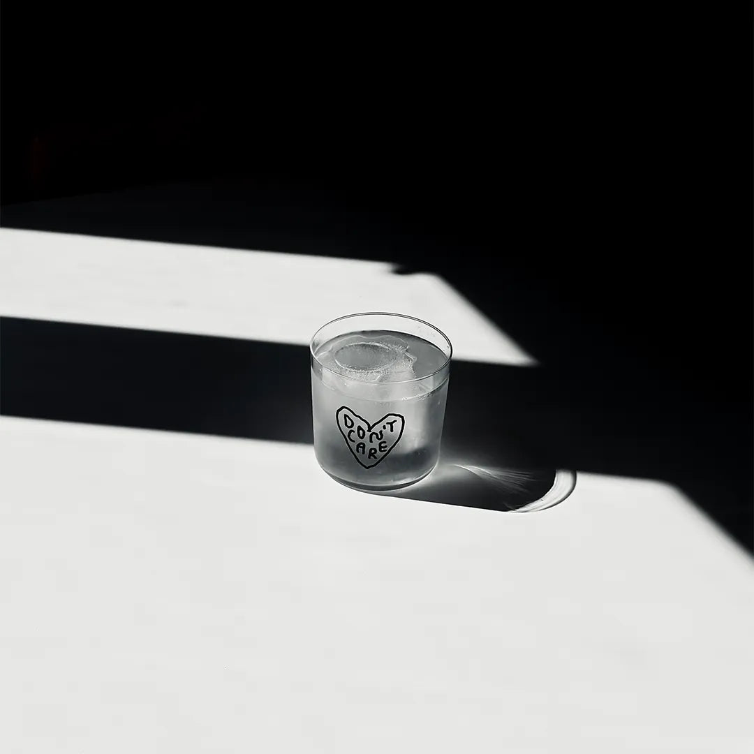 Transparentes Medium Designer Glas mit schwarzem Schriftzug "Don't Care" geschrieben in einem Herz. Das Glas steht in einem Lichtkegel, viel Schatten im Hintergrund, auf einem weißen Tisch.