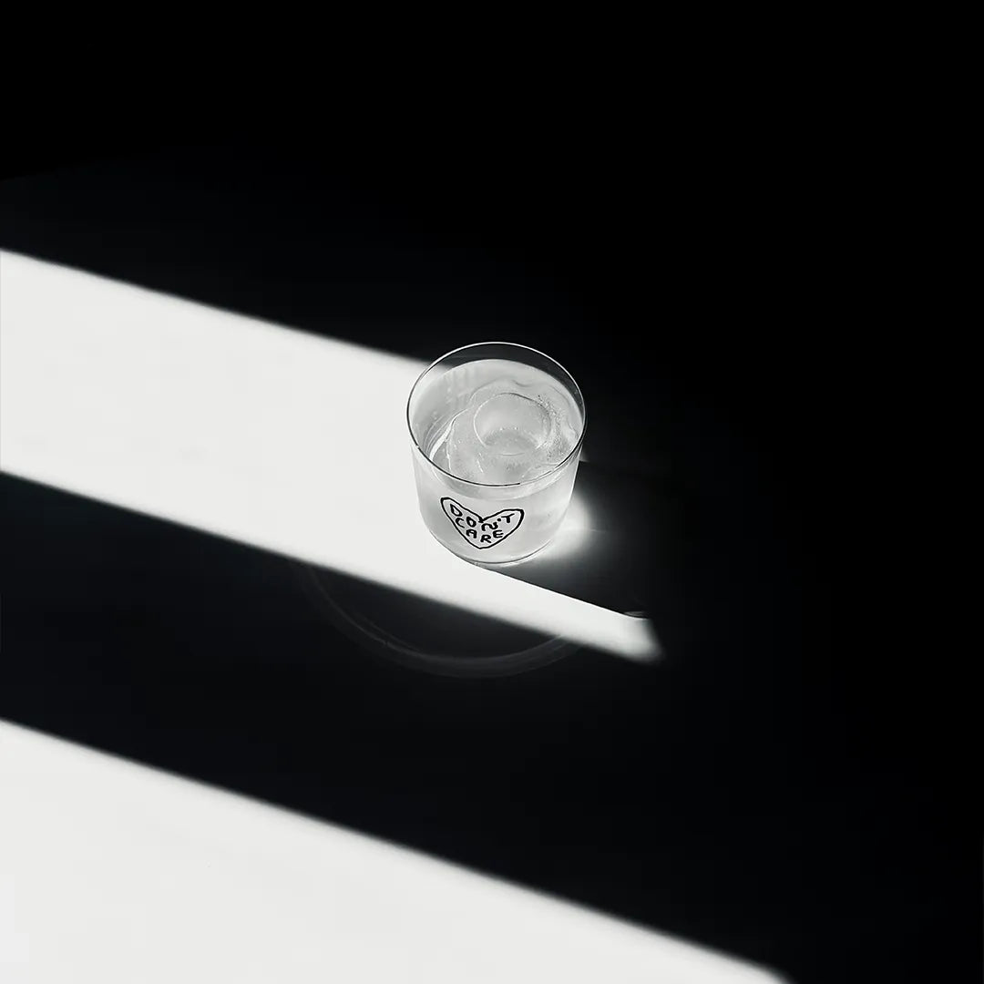 Transparentes Medium Designer Glas mit schwarzem Schriftzug "Don't Care" geschrieben in einem Herz. Das Glas steht in einem Lichtkegel, viel Schatten im Hintergrund, auf einem weißen Tisch.