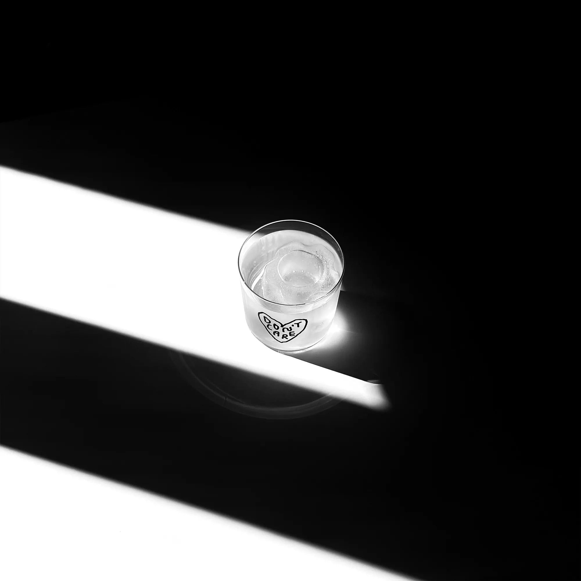 Transparentes Medium Designer Glas mit schwarzem Schriftzug "Don't Care" geschrieben in einem Herz. Das Glas steht in einem Lichtkegel, viel Schatten im Hintergrund, auf einem weißen Tisch.