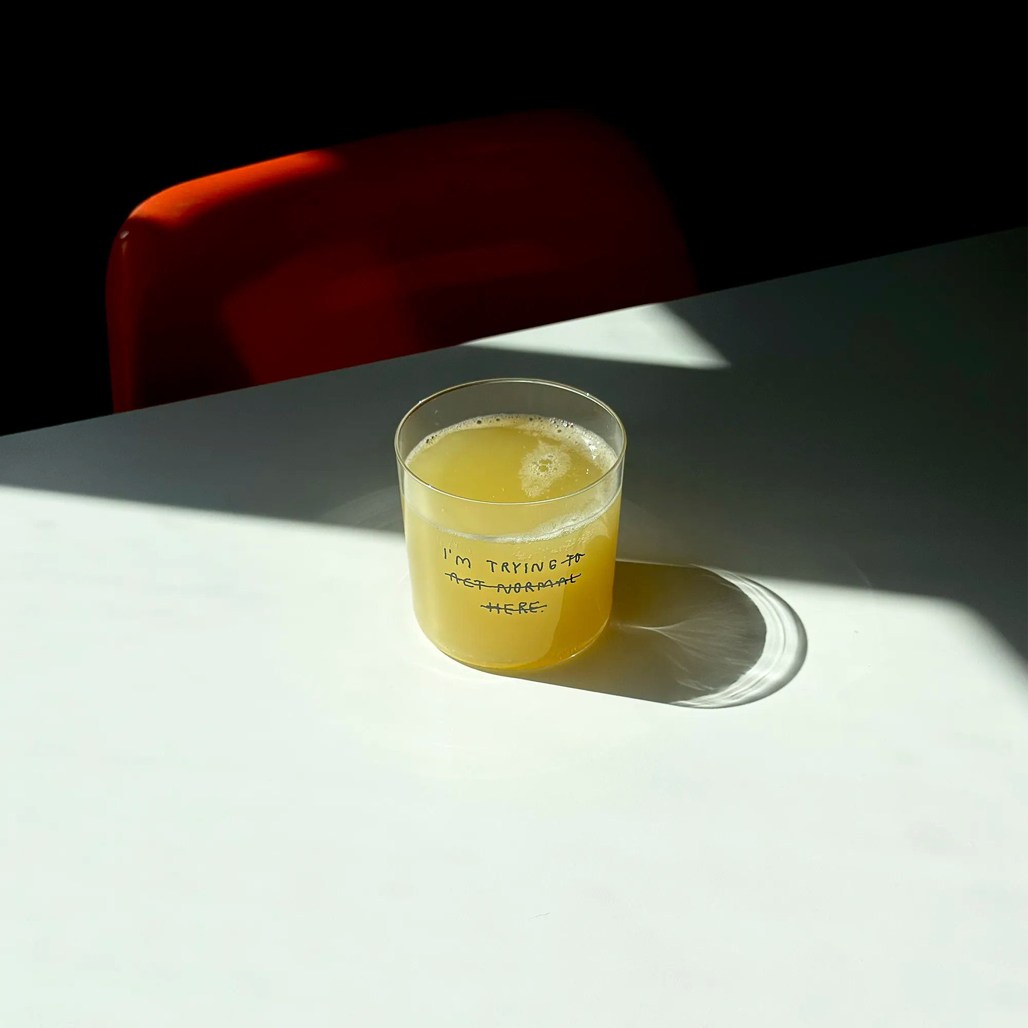 Transparentes Medium Designer Glas mit schwarzem Schriftzug "i'm trying to act normal here" mit dünnen Wänden und Boden. Dabei ist das "to act normal here durchgestrichen, so dass lediglich die Aussage "I'm trying" steht.

Das Glas ist mit einer gelben Flüssigkeit befüllt und Eiswürfeln. Es steht auf einem weißen Tisch und wirft einen schatten.