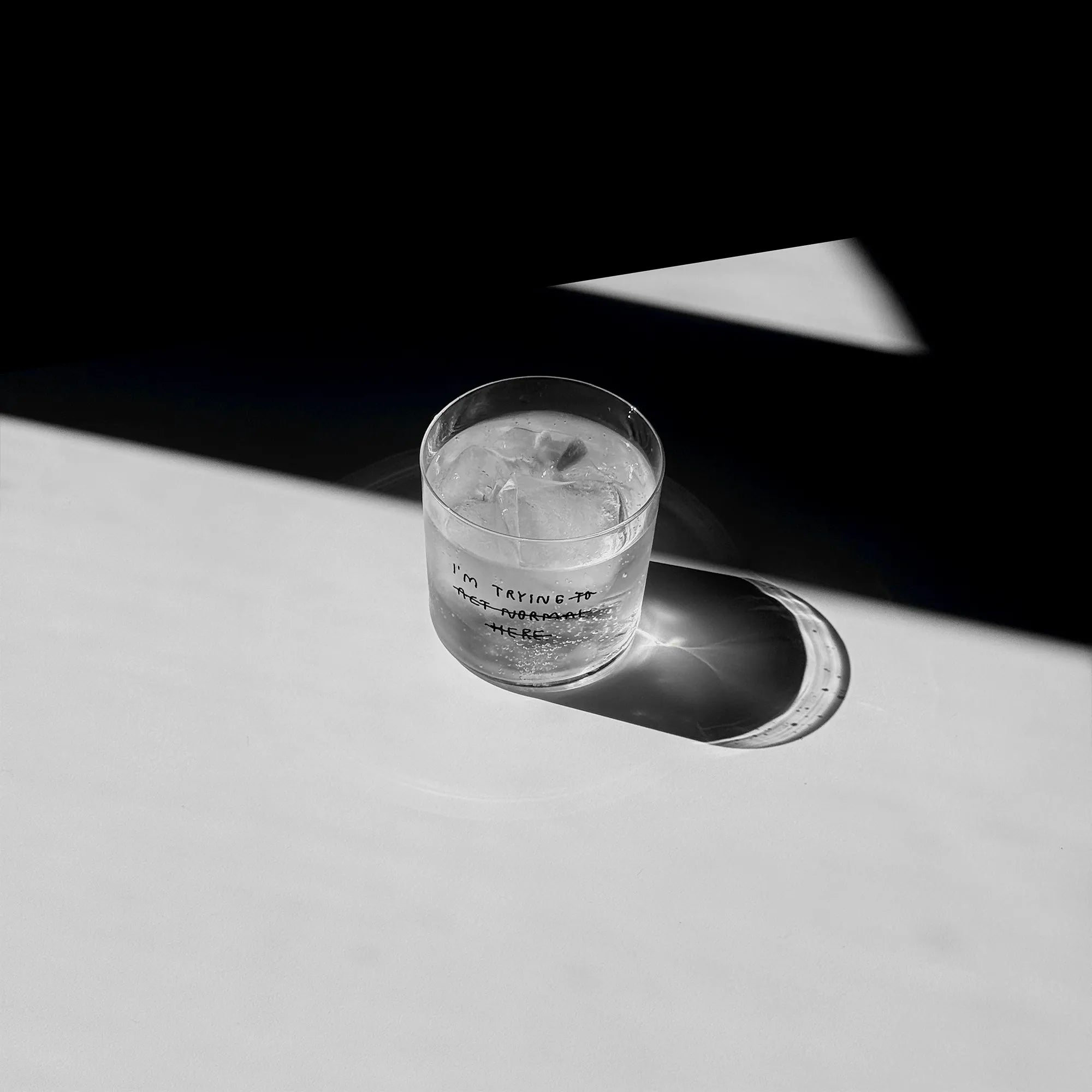 Transparentes Medium Designer Glas mit schwarzem Schriftzug "i'm trying to act normal here" mit dünnen Wänden und Boden. Dabei ist das "to act normal here durchgestrichen, so dass lediglich die Aussage "I'm trying" steht.

Das Glas ist mit einer transparenten Flüssigkeit befüllt und Eiswürfeln. Es steht auf einem weißen Tisch und wirft einen schatten.