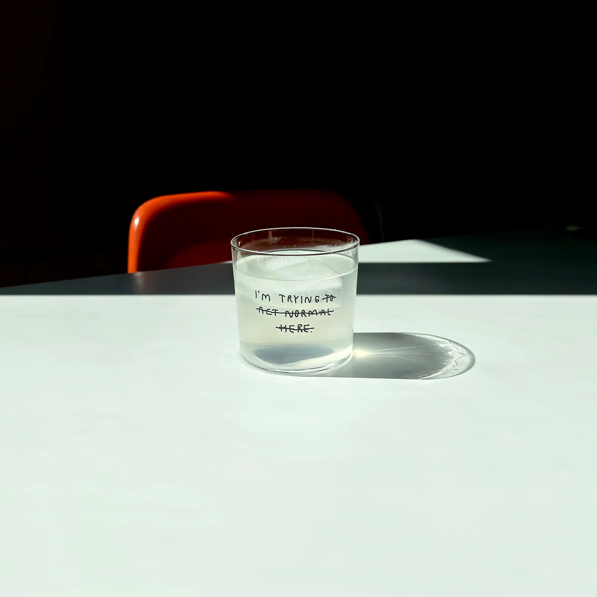 Transparentes Medium Designer Glas mit schwarzem Schriftzug "i'm trying to act normal here" mit dünnen Wänden und Boden. Dabei ist das "to act normal here durchgestrichen, so dass lediglich die Aussage "I'm trying" steht.

Das Glas ist mit einer transparenten Flüssigkeit befüllt und Eiswürfeln. Es steht auf einem weißen Tisch und wirft einen schatten.