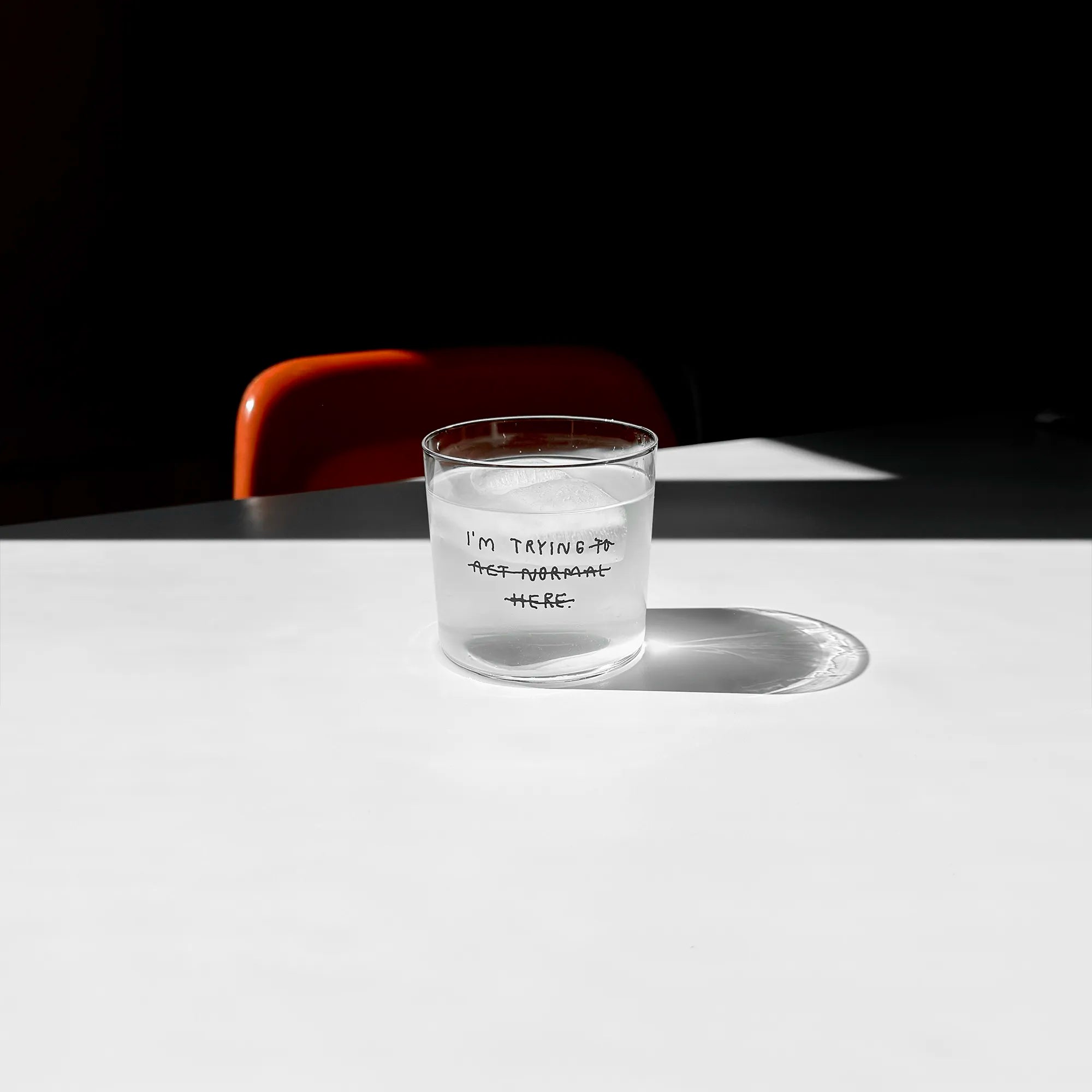 Transparentes Medium Designer Glas mit schwarzem Schriftzug "i'm trying to act normal here" mit dünnen Wänden und Boden. Dabei ist das "to act normal here durchgestrichen, so dass lediglich die Aussage "I'm trying" steht.

Das Glas ist mit einer transparenten Flüssigkeit befüllt und Eiswürfeln. Es steht auf einem weißen Tisch und wirft einen schatten.