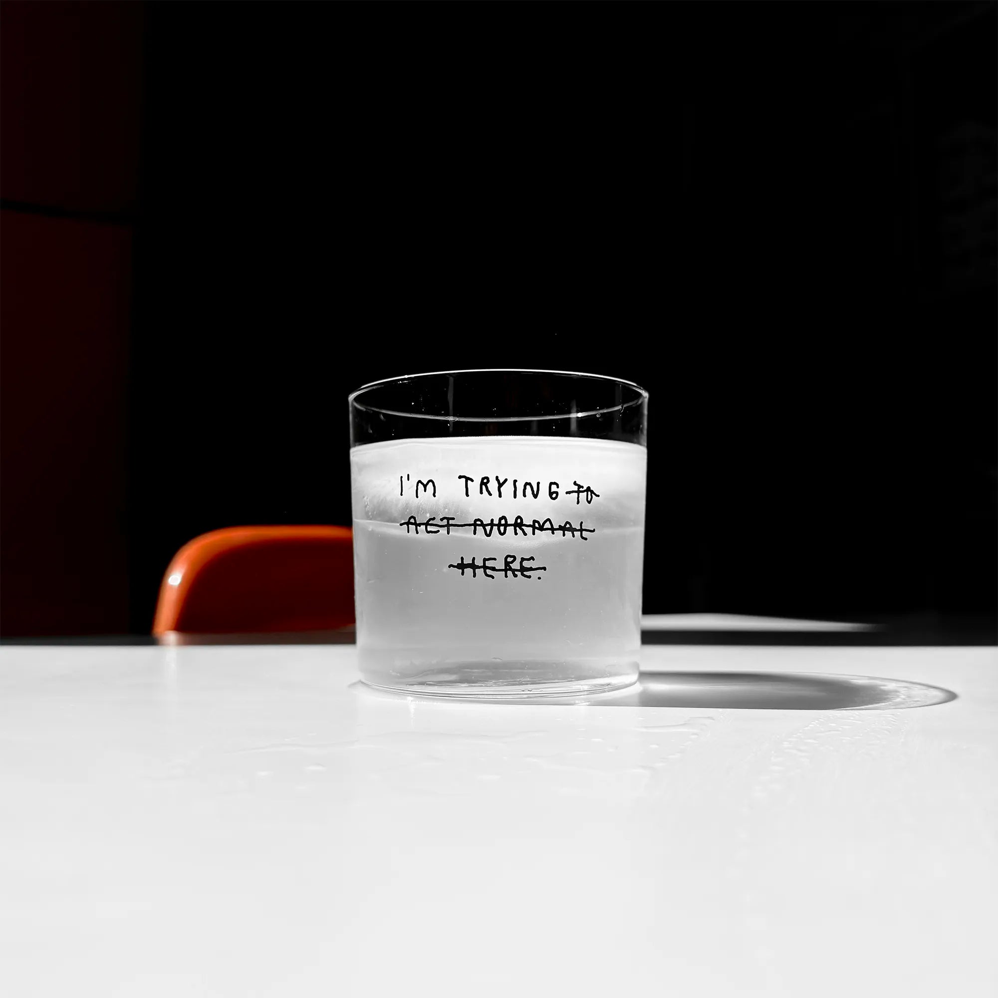 Transparentes Medium Designer Glas mit schwarzem Schriftzug "i'm trying to act normal here" mit dünnen Wänden und Boden. Dabei ist das "to act normal here durchgestrichen, so dass lediglich die Aussage "I'm trying" steht.

Das Glas ist mit einer transparenten Flüssigkeit befüllt und Eiswürfeln. Es steht auf einem weißen Tisch und wirft einen schatten.