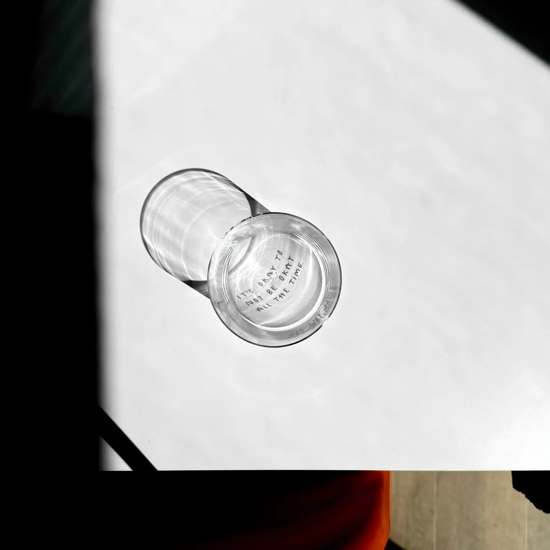 Transparentes Medium Designer Glas mit schwarzem Schriftzug "It's okay to not be okay all the time" mit dünnen Wänden und Boden. 

Das Glas ist unbefüllt und ein Foto von oben geschossen so dass man das Motiv nur als Schattenwurf im Glas sieht. 