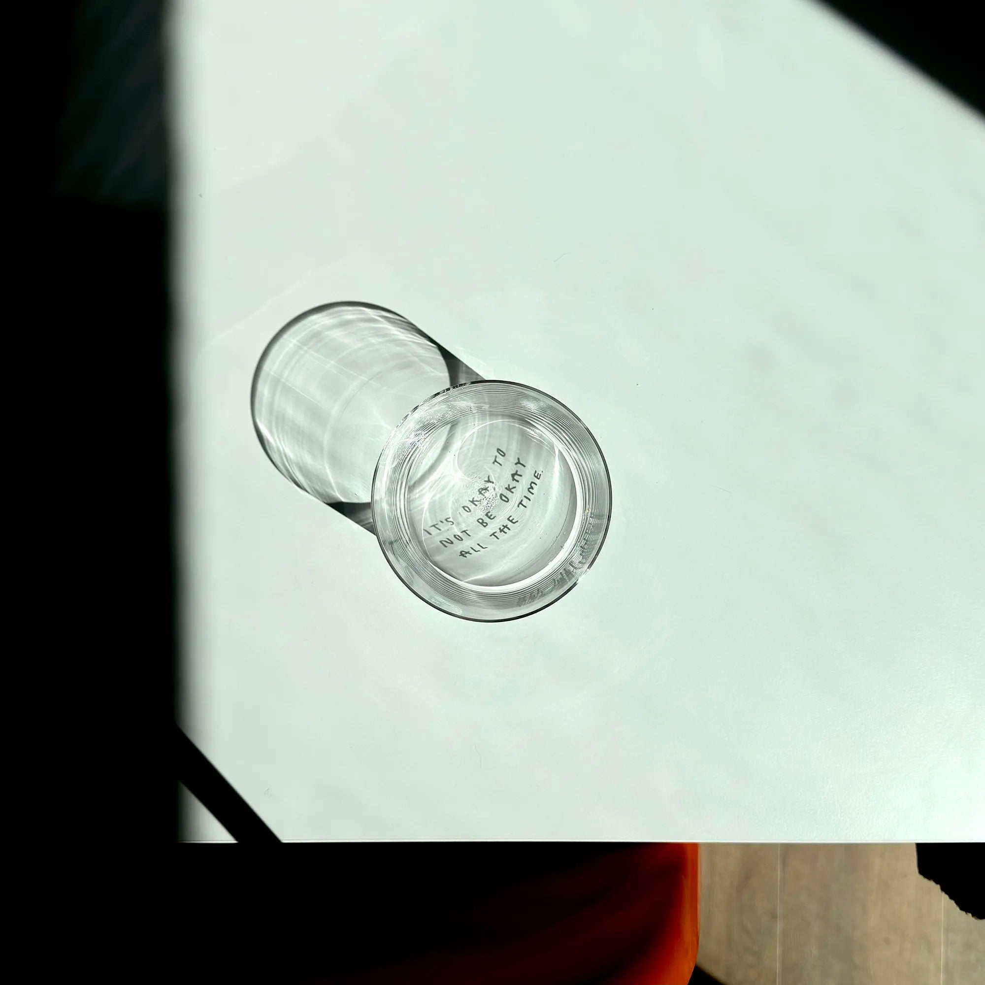 Transparentes Medium Designer Glas mit schwarzem Schriftzug "It's okay to not be okay all the time" mit dünnen Wänden und Boden. 

Das Glas ist unbefüllt und ein Foto von oben geschossen so dass man das Motiv nur als Schattenwurf im Glas sieht. 