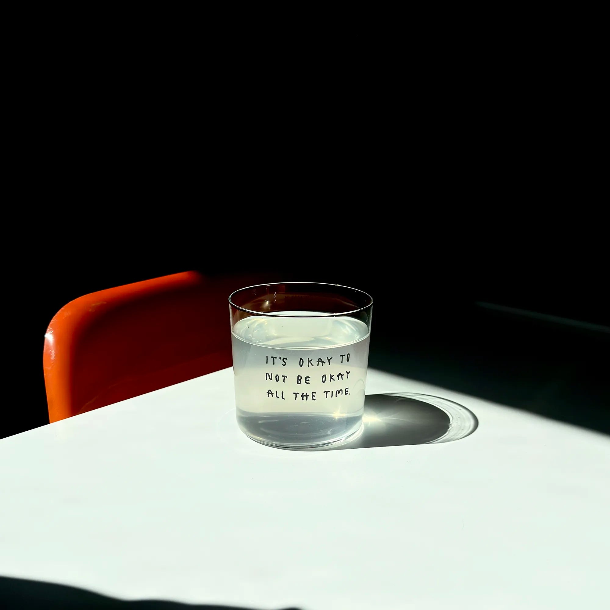 Transparentes Medium Designer Glas mit schwarzem Schriftzug "It's okay to not be okay all the time" mit dünnen Wänden und Boden. 

Das Glas ist mit einer transparenten Flüssigkeit befüllt. Es steht auf einem weißen Tisch und wirft einen schatten. Daneben steht ein orangener Stuhl.