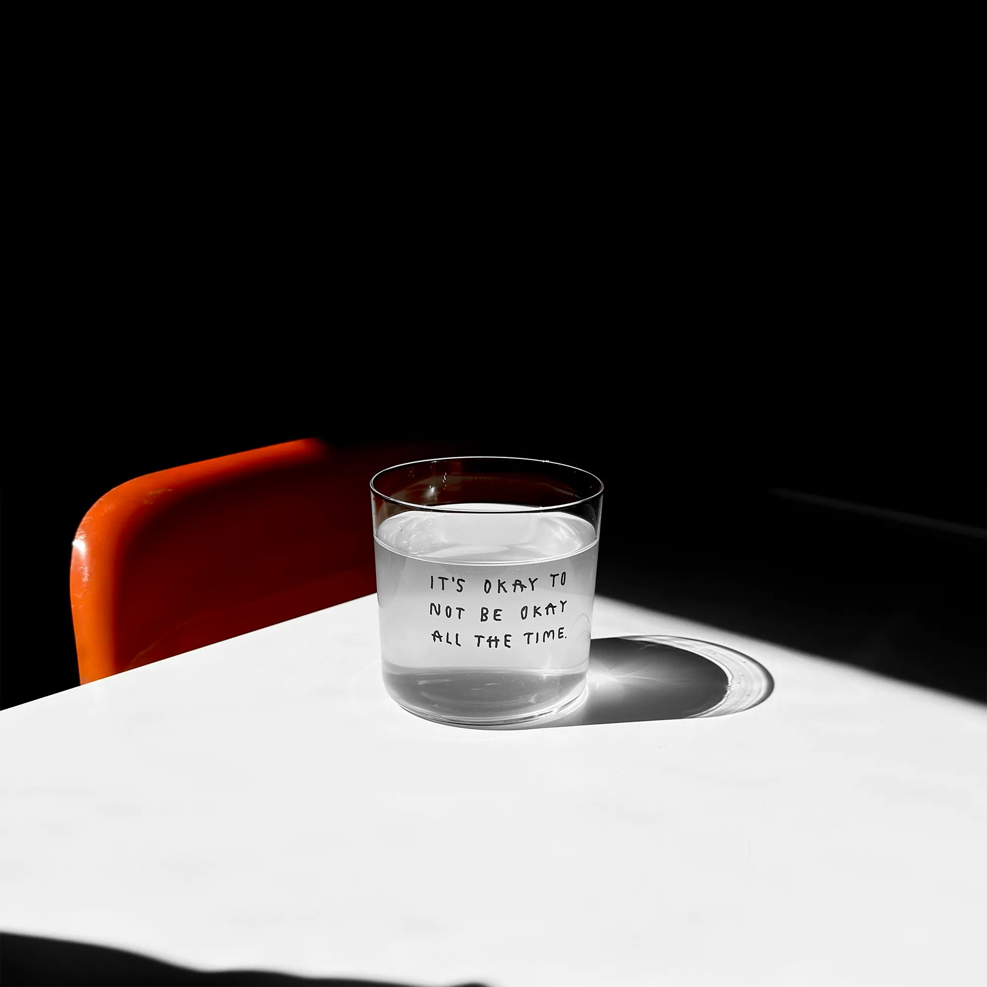 Transparentes Medium Designer Glas mit schwarzem Schriftzug "It's okay to not be okay all the time" mit dünnen Wänden und Boden. 

Das Glas ist mit einer transparenten Flüssigkeit befüllt. Es steht auf einem weißen Tisch und wirft einen schatten. Daneben steht ein orangener Stuhl.