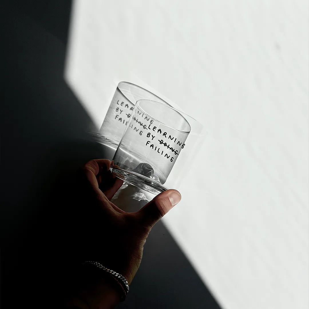 Klares, zylindrisches Trinkglas mit schwarzem, handschriftlich wirkendem Text. Darauf steht „Learning by doing“, wobei das Wort „doing“ durchgestrichen ist und darunter „failing“ steht, sodass der Satz humorvoll zu „Learning by failing“ geändert wird.

Das Glas liegt auf einem weißen Tisch, zur Hälfte in Schatten und wird von einer Hand am unteren Rand gehalten.