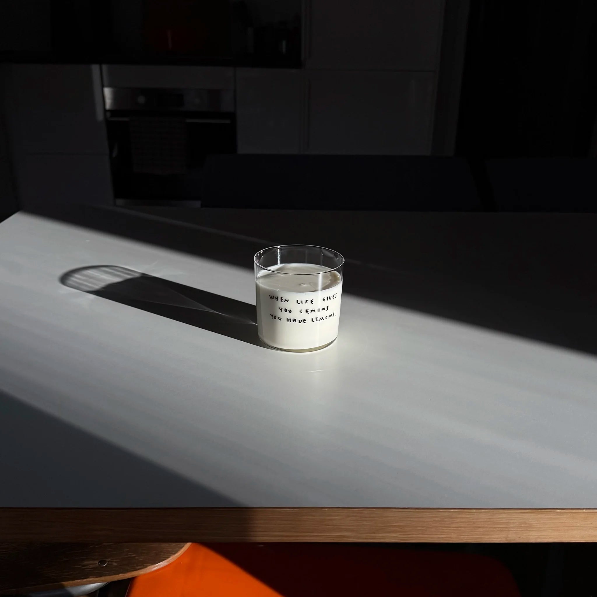 Transparentes Medium Designer Glas mit schwarzem Schriftzug "when life gives you lemons" mit dünnen Wänden und Boden. Das Glas steht im Licht auf einem weißen Tisch und wirft einen weiten Schatten. Das Glas ist mit Milch befüllt. 