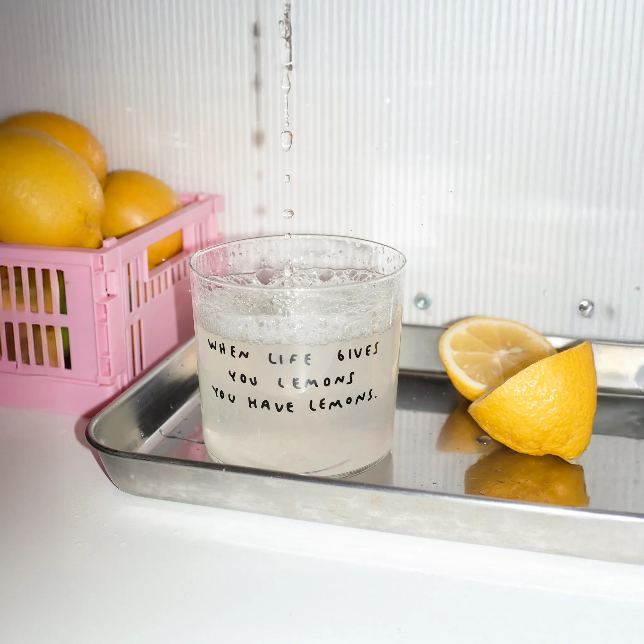 Transparentes Medium Designer Glas mit schwarzem Schriftzug "when life gives you lemons" mit dünnen Wänden und Boden.
Das Glas wird mit einer Flüssigkeit befüllt, daneben liegen Zitronen. Das Glas steht auf einem Aluminium-Brett.