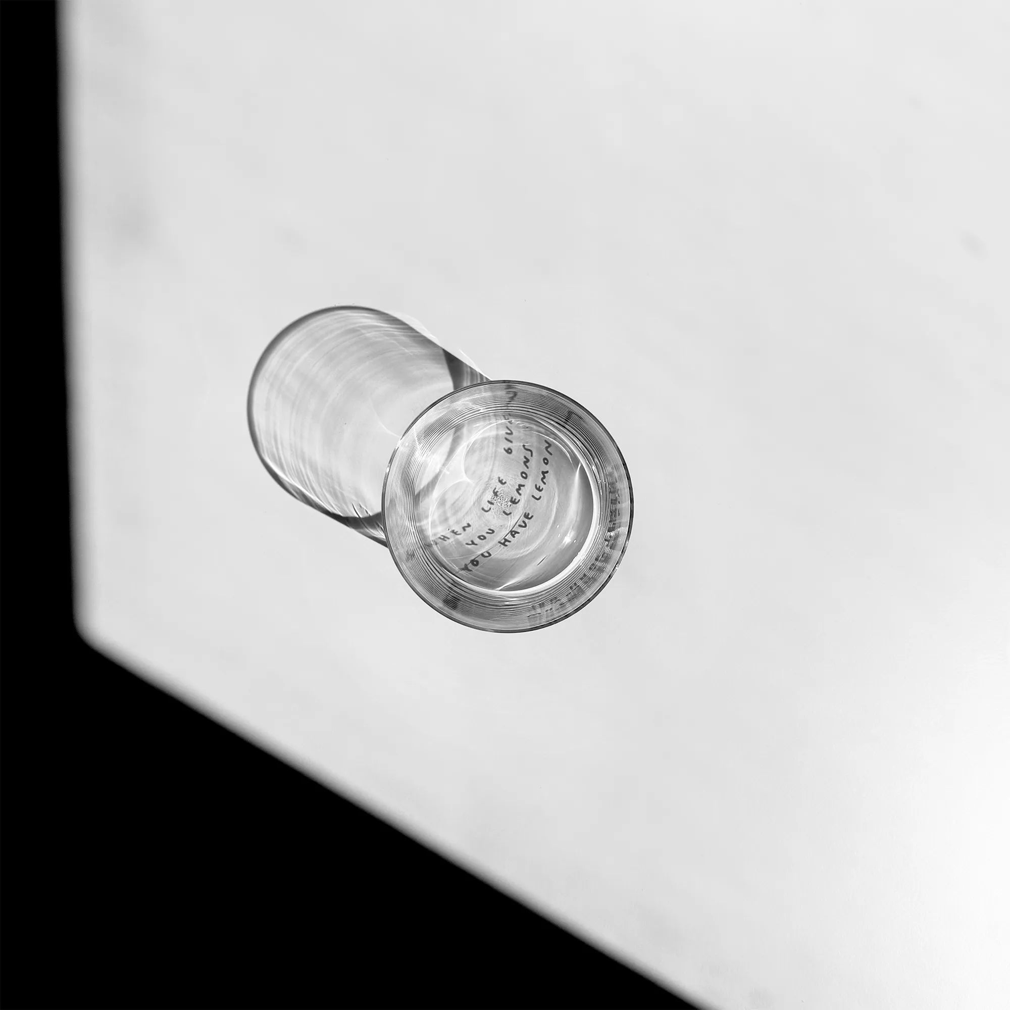 Transparentes Medium Designer Glas mit schwarzem Schriftzug "when life gives you lemons" mit dünnen Wänden und Boden. Das Glas steht im Licht auf einem weißen Tisch. Der Rest des Bildes ist dunkel in Schatten gefallen.