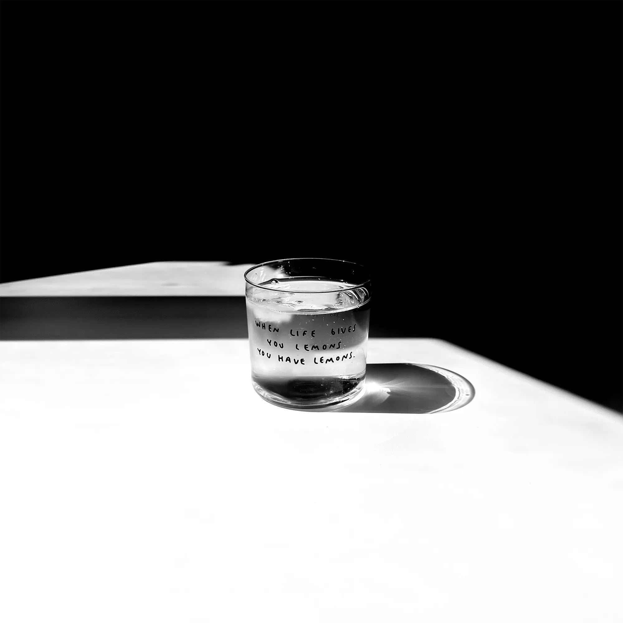 Transparentes Medium Designer Glas mit schwarzem Schriftzug "when life gives you lemons" mit dünnen Wänden und Boden. Das Glas steht im Licht auf einem weißen Tisch. Der Rest des Bildes ist dunkel in Schatten gefallen.

Das Glas ist mit einer transparenten Flüssigkeit befüllt.