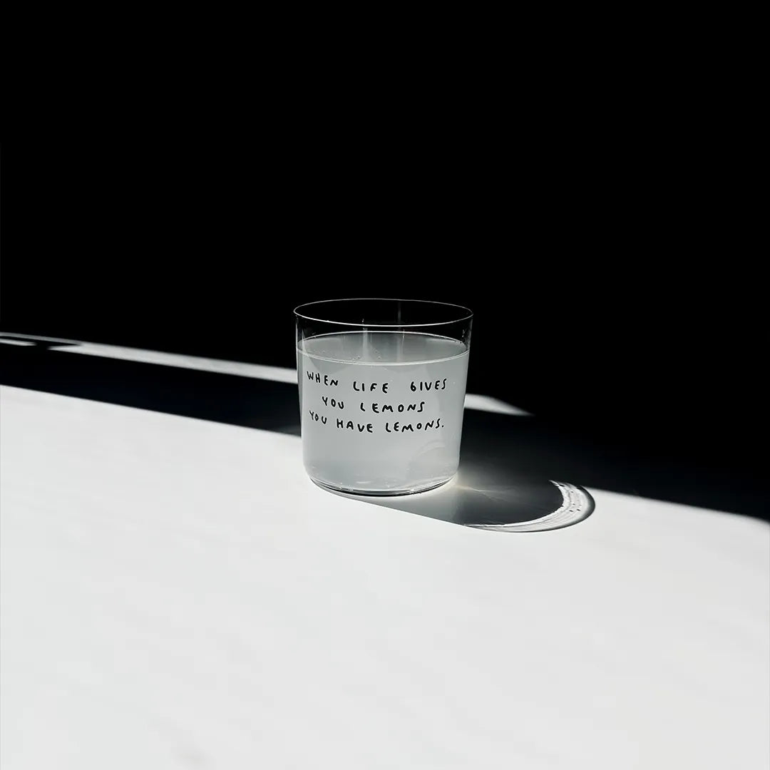 Transparentes Medium Designer Glas mit schwarzem Schriftzug "when life gives you lemons" mit dünnen Wänden und Boden. Das Glas steht im Licht auf einem weißen Tisch. Der Rest des Bildes ist dunkel in Schatten gefallen.

Das Glas ist mit einer hellen Flüssigkeit befüllt.
